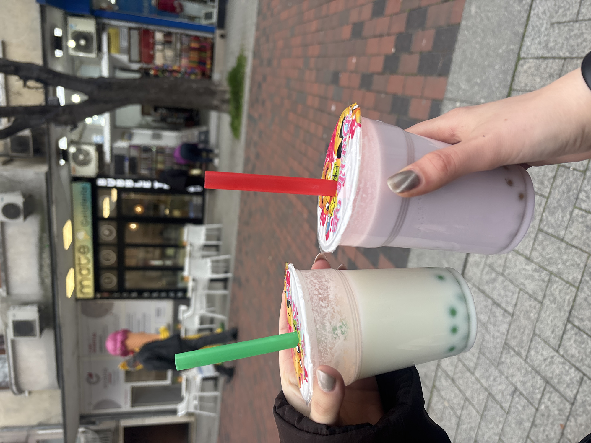 The image shows two hands holding bubble tea drinks. One drink is lavender-colored with a red straw, the other is milky-green with a green straw. The background shows a sidewalk with red brick and grey stone. Across the street is a building with white chairs and tables outside, and a sign that says "mate | cafetterie". A person with a pink hat is visible in the background. Based on the sign, the location is most likely a cafe called "mate cafetterie". Without additional information, it is not possible to pinpoint the location more precisely in Varna.