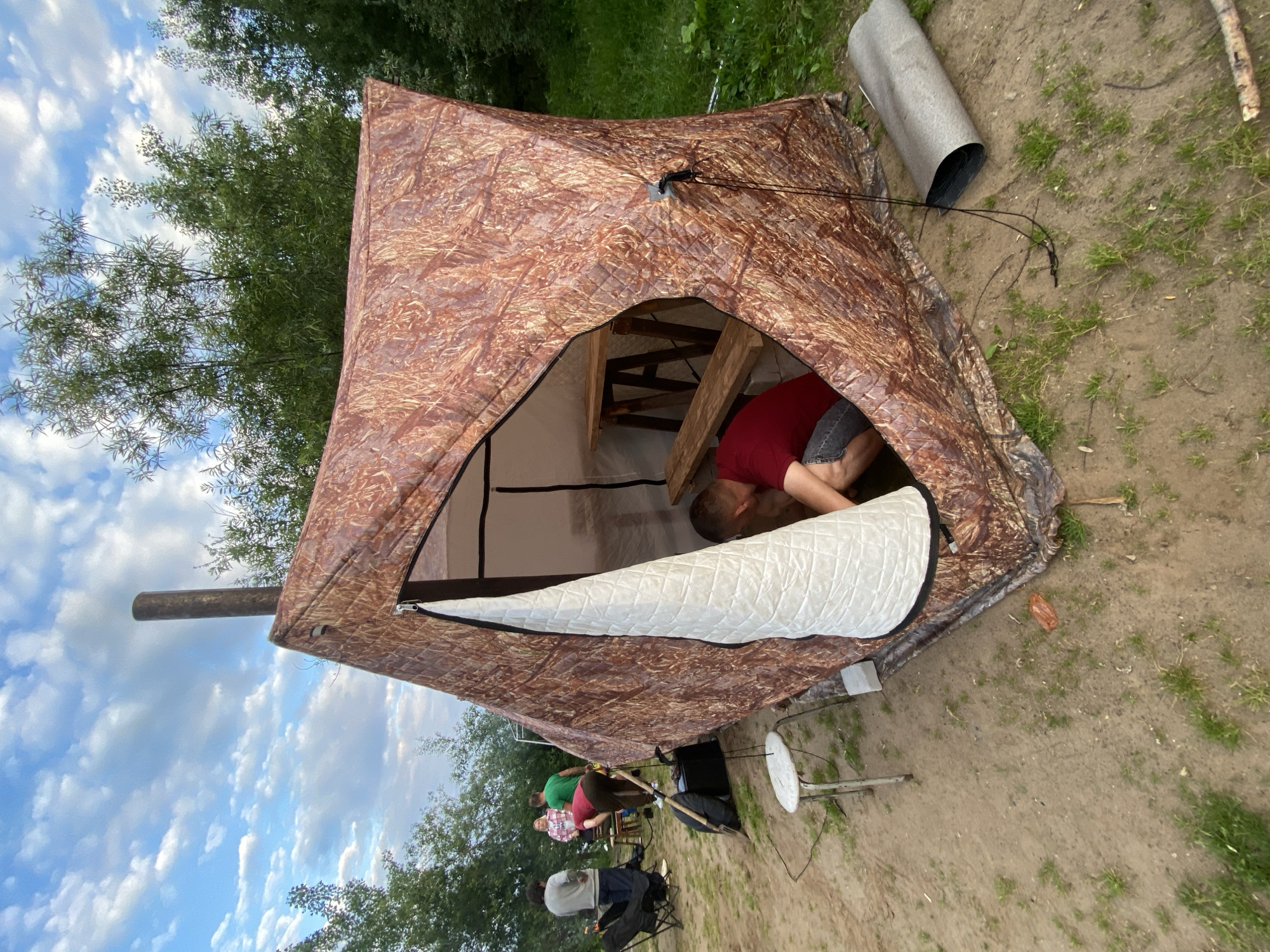 The image depicts a campsite in Noviy, Russia, under a cloudy sky. In the foreground, a large, camouflage-patterned tent with a chimney is pitched. A man in a red shirt and denim shorts is partially visible inside the tent, tending to something on the ground. Outside the tent, on the left, three people are standing. One person is wearing a grey sweater and jeans, another a pink and white checkered shirt, and the third a green t-shirt. A black camping chair is set up nearby. The ground is a mix of dirt and sparse grass, with some fallen leaves and twigs. To the right of the tent, a rolled-up grey mat lies on the ground. In the background, dense green trees line the horizon.