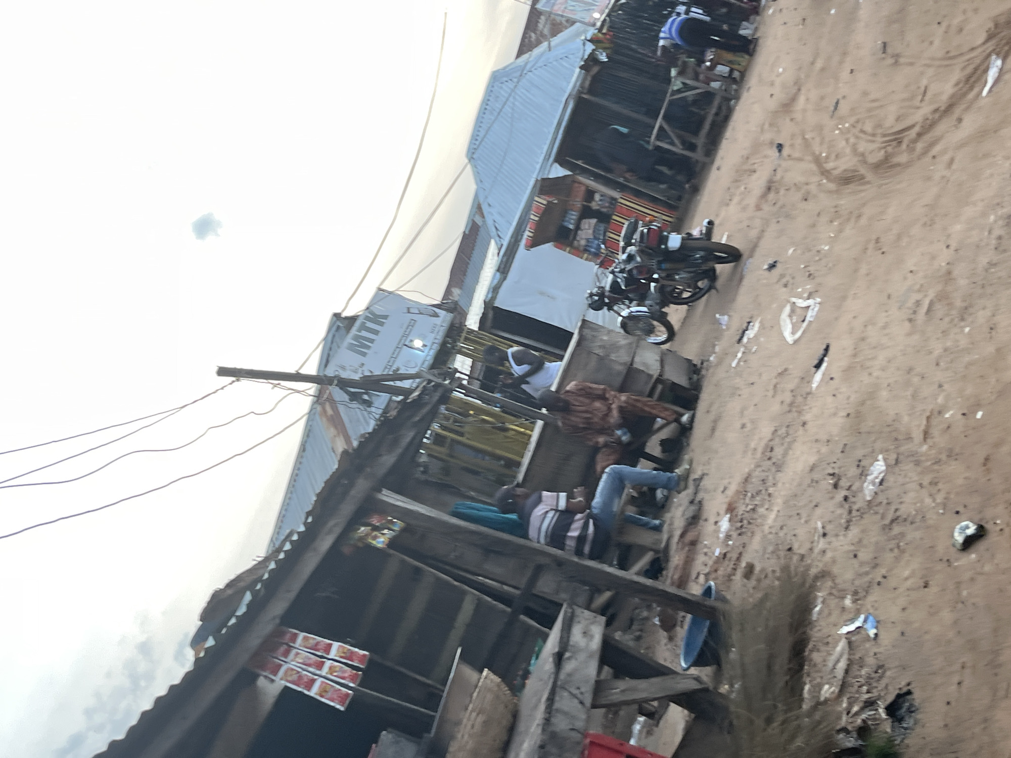 The image depicts a bustling outdoor market scene. Several individuals are present, some seated and others standing, amidst temporary structures and stalls. A motorcycle is parked prominently in the foreground, suggesting transportation is a key element of the scene. The setting appears to be a dirt-floored area, possibly a roadside market or a village square, with a mix of corrugated metal and wooden structures.
One person is wearing a striped shirt and jeans, seated on what appears to be a wooden platform. Another individual in the background is wearing a patterned shirt and jeans. A third person is visible in a blue top and jeans. The overall impression is one of everyday activity and commerce.
The time of day appears to be late afternoon or early evening, given the soft, diffused light. The sky is visible, with some clouds, but no strong shadows are cast, indicating overcast or twilight conditions. There are no specific location cues to pinpoint the exact geographical area, but the rustic nature of the market suggests a developing region.
A white truck with the letters "MTK" emblazoned on its side is visible in the middle ground, adding to the sense of a commercial hub. The ground is littered with debris and some plastic bags, typical of a busy market environment. The image is oriented sideways, making it appear as if the scene is tilted.
The activity seems to be a moment of rest or interaction among the people in the market. There are no overt signs of distress or urgency; rather, the scene conveys a sense of ordinary life unfolding. The presence of multiple people and the parked motorcycle suggest a place where people gather and transact business.