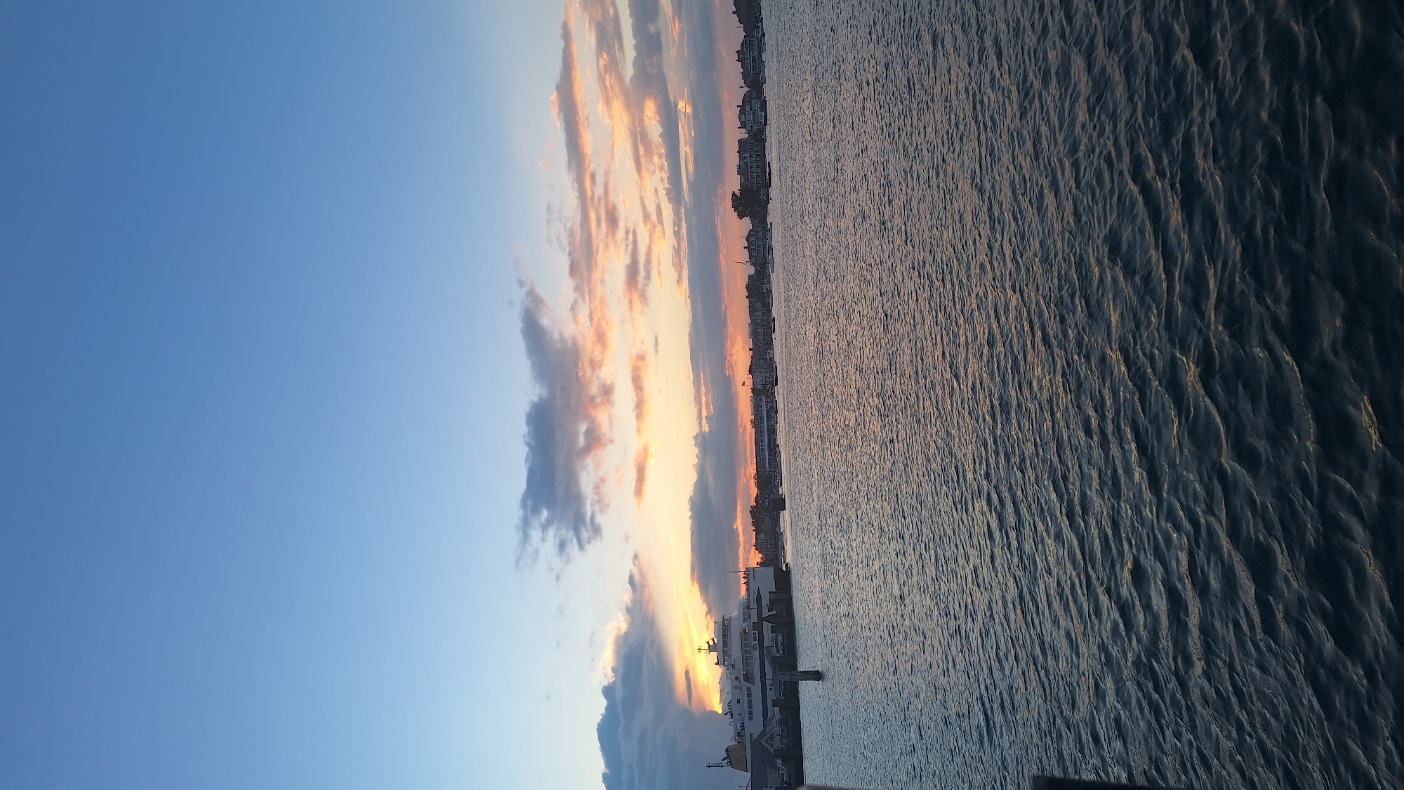 This image captures a serene sunset over the waters of Nantucket, Massachusetts. The sky is a gradient of clear blue at the top, transitioning to soft hues of pink, orange, and yellow as it meets the horizon, illuminated by the setting sun behind scattered clouds. The water in the foreground reflects the colors of the sky, creating a shimmering, textured surface.
On the left side of the frame, a large white ferry is docked, its structure clearly visible. Several vehicles are parked on the ferry and on the dock adjacent to it. A tall, dark, cylindrical marker stands in the water near the ferry. In the background, along the waterfront, a line of buildings with distinct architecture, characteristic of Nantucket, stretches across the horizon. Numerous sailboats and smaller vessels are anchored or moored in the harbor. The overall atmosphere is tranquil, conveying the peaceful end of a day in this picturesque coastal town.