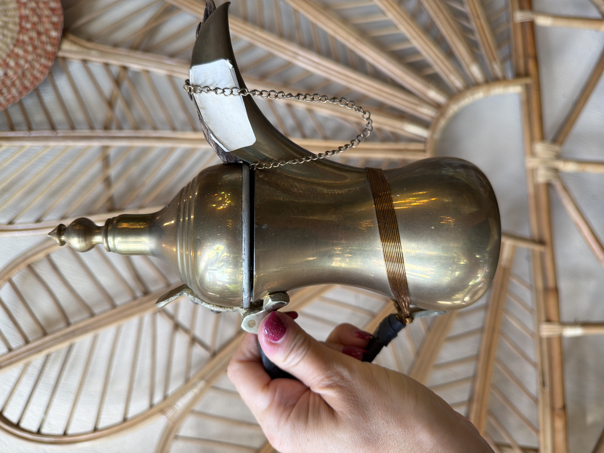 A hand with red nail polish holds the handle of a traditional brass dallah, a type of Arabic coffee pot, against a backdrop of light-colored woven rattan. The dallah features a long, thin spout with a small chain attached to its lid, and a decorative band of coiled wire around its body. The rattan background is arranged in a fan-like pattern. This object is likely located in Üsküdar, Turkey, a district known for its cultural heritage and markets where such items might be found.