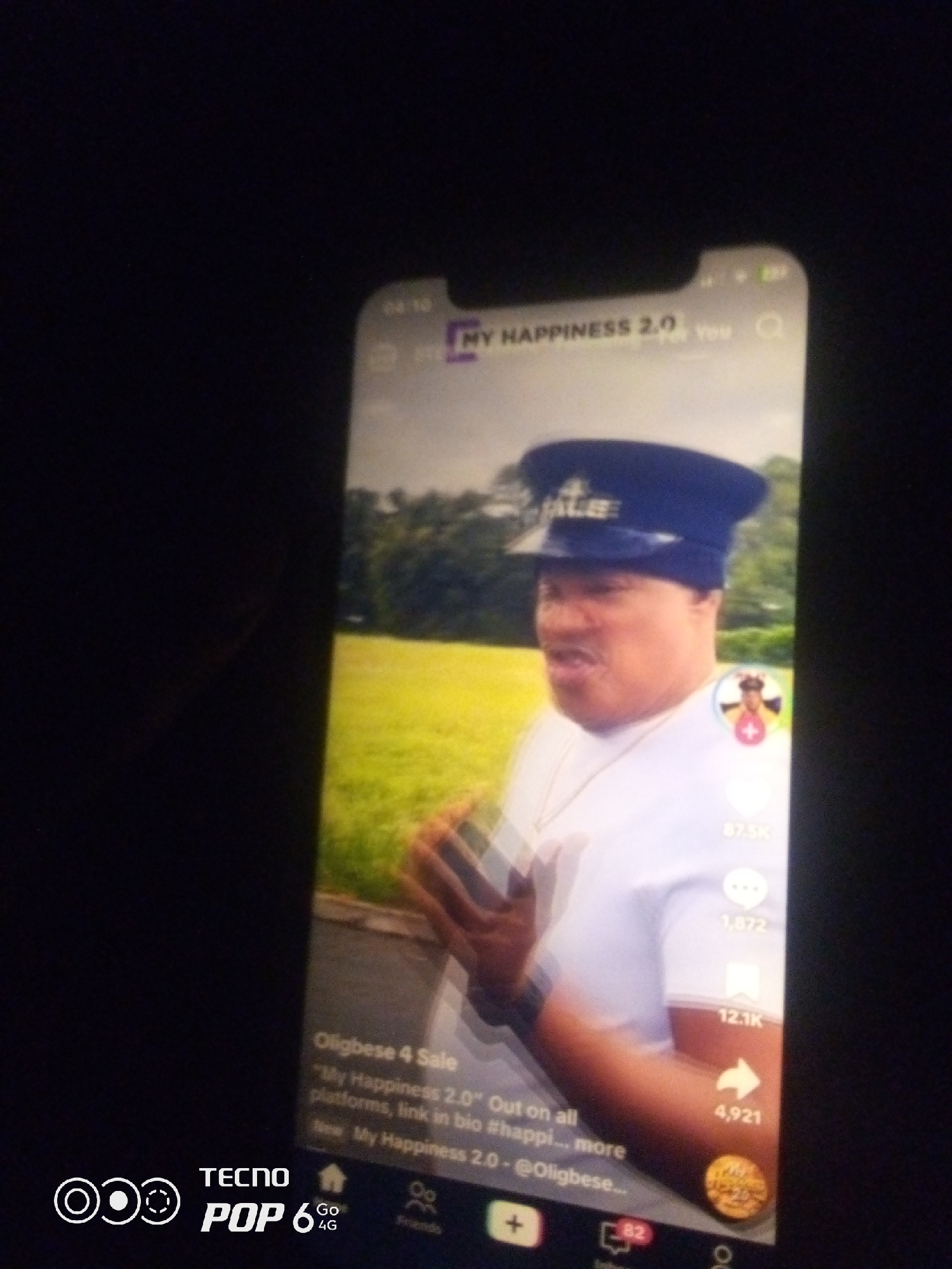 A dimly lit, vertical, close-up photograph shows a person wearing a navy blue peaked cap and a white t-shirt. The person is positioned in the center of the frame, facing slightly to the left. The background consists of a blurred green field and trees. The image appears to be a screenshot from a social media application, indicated by icons and text overlays. On the right side of the screen, there are engagement metrics such as "87.5K," "1,872," "12.1K," and "4,921," along with a red heart icon and a comment icon. At the bottom of the screen, there are navigation icons, including a home icon, a message icon with "82" next to it, and a profile icon. There is also a logo for "TECNO POP 6 4G" in the bottom left corner. The caption below the video reads "Oligbese 4 Sale" and "My Happiness 2.0" Out on all platforms, link in bio #happi... more New My Happiness 2.0 - @Oligbese_...". The image does not provide any specific details about the location in the city of Howayi, Nigeria.