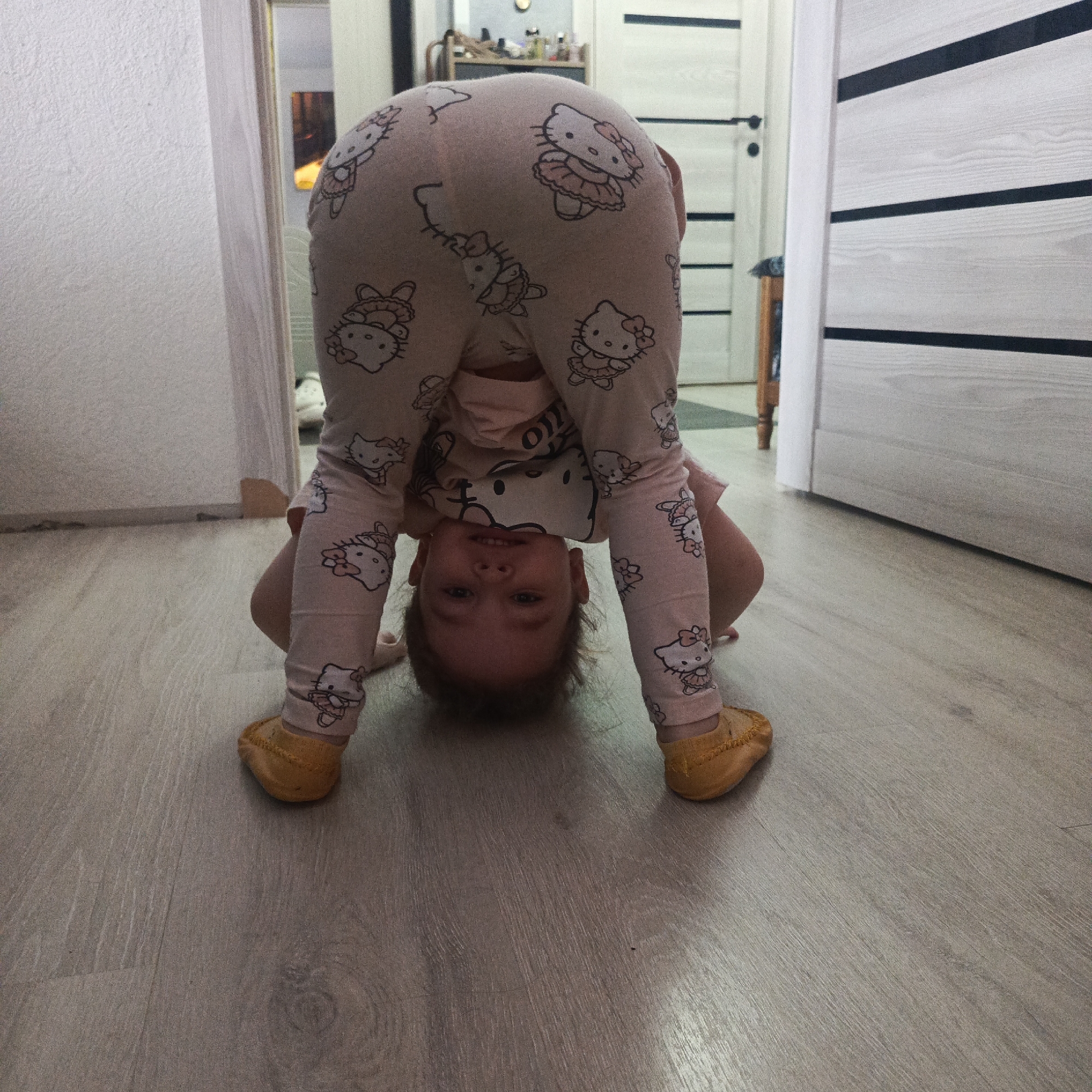 A young child in pink Hello Kitty pajamas and yellow shoes is upside down, supported by their hands on a light-colored wood-grain floor. The child's face is visible, looking towards the camera. The location appears to be an interior space, possibly a home, with doorways and a white and black striped door in the background. Based on the prompt, the location is presumed to be in Марковский, Russia, although this cannot be confirmed visually.