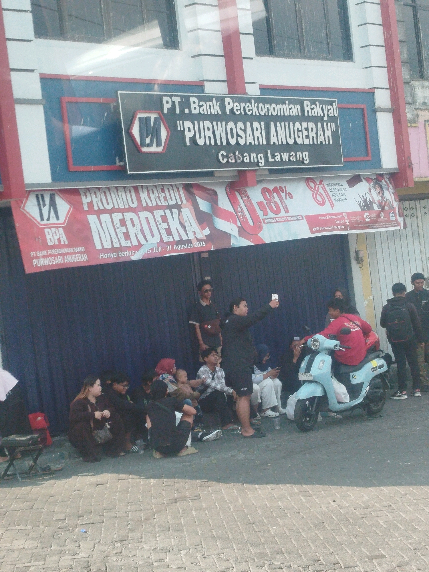 The image depicts the exterior of a bank branch in Lawang, Indonesia. The bank's signage reads "PT. Bank Perekonomian Rakyat 'PURWOSARI ANUGERAH' Cabang Lawang." Below this, a red banner advertises "PROMO KREDIT MERDEKA" with details indicating it runs from July 15 to August 31, 2026.
In the foreground, a group of people are gathered on a paved area, some sitting and some standing. Several individuals are looking at their phones. A light blue scooter is parked prominently in the center, with one person seated on it and another standing nearby. Other individuals are visible in the background, some near the entrance of the bank. The environment suggests a public outdoor space adjacent to a commercial building.