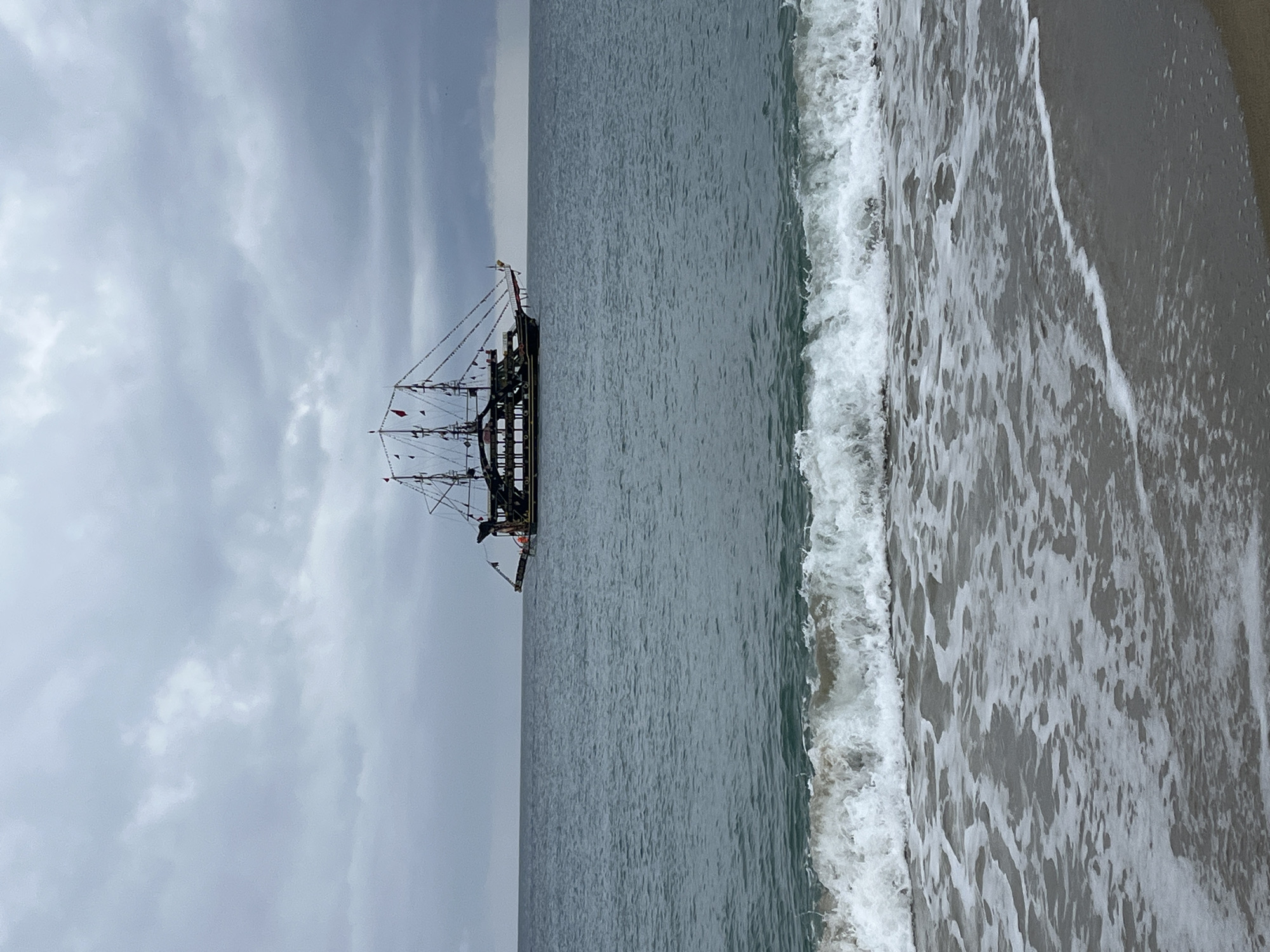 The image depicts a beach scene in Alanya, Turkey. A large, dark-colored pirate ship is anchored off the coast. The sky is overcast. Waves are crashing on the shore. The beach appears to be made of dark sand.