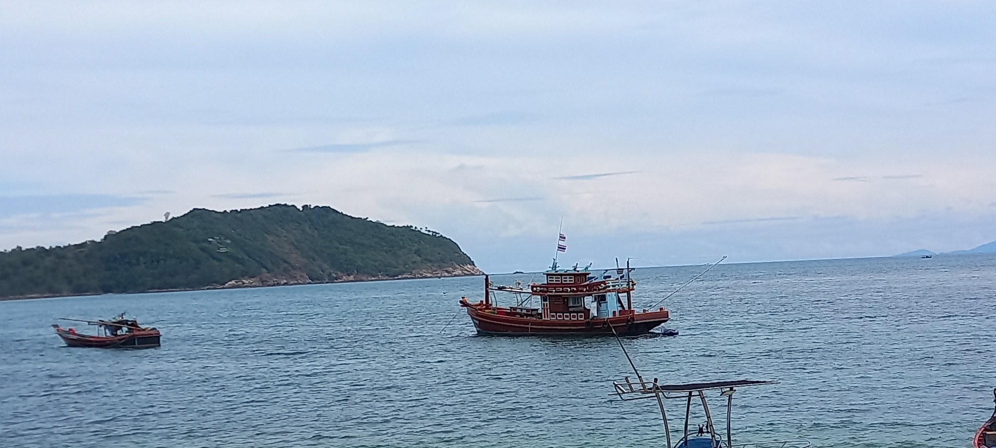 The image depicts a coastal scene near Baan Chaloklum, Thailand. In the foreground, the ocean water displays subtle ripples. Two wooden fishing boats are visible, one in the near-center of the frame and another smaller one to the left. The primary boat, larger and painted in shades of red, carries a Thai flag. An island covered in green vegetation forms the backdrop on the left side of the frame. The sky is overcast with muted white and grey clouds. Additional boat structure visible in the lower right corner.