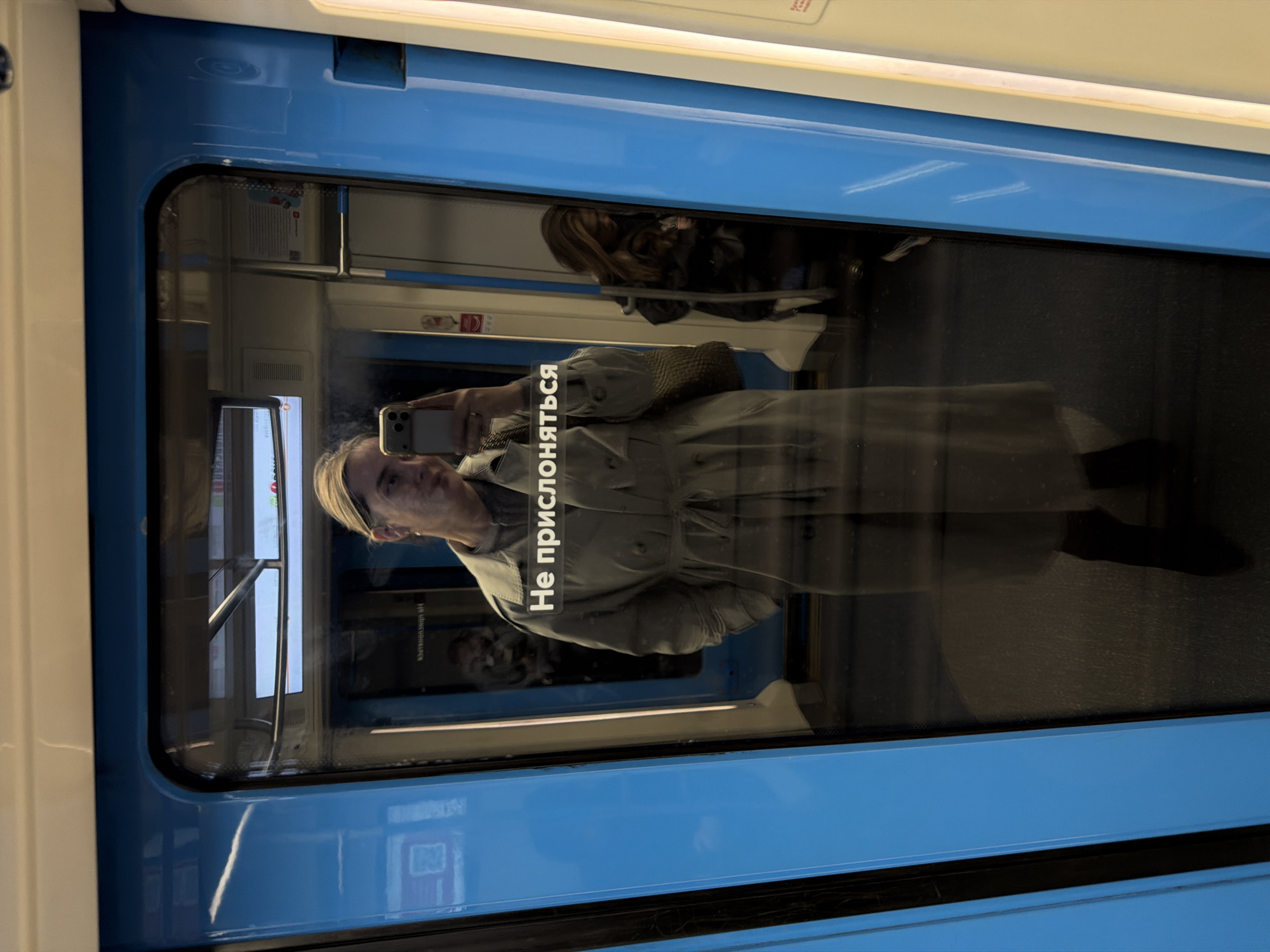 The image shows a reflection in a train window, likely in the Moscow Metro, Russia. The reflection features a person taking a selfie with a smartphone. The person is wearing a light-colored trench coat. The train window has a sticker that reads "Не прислоняться" which means "Do not lean." Other passengers can be seen in the background through the window. The train car itself is painted in blue and white.