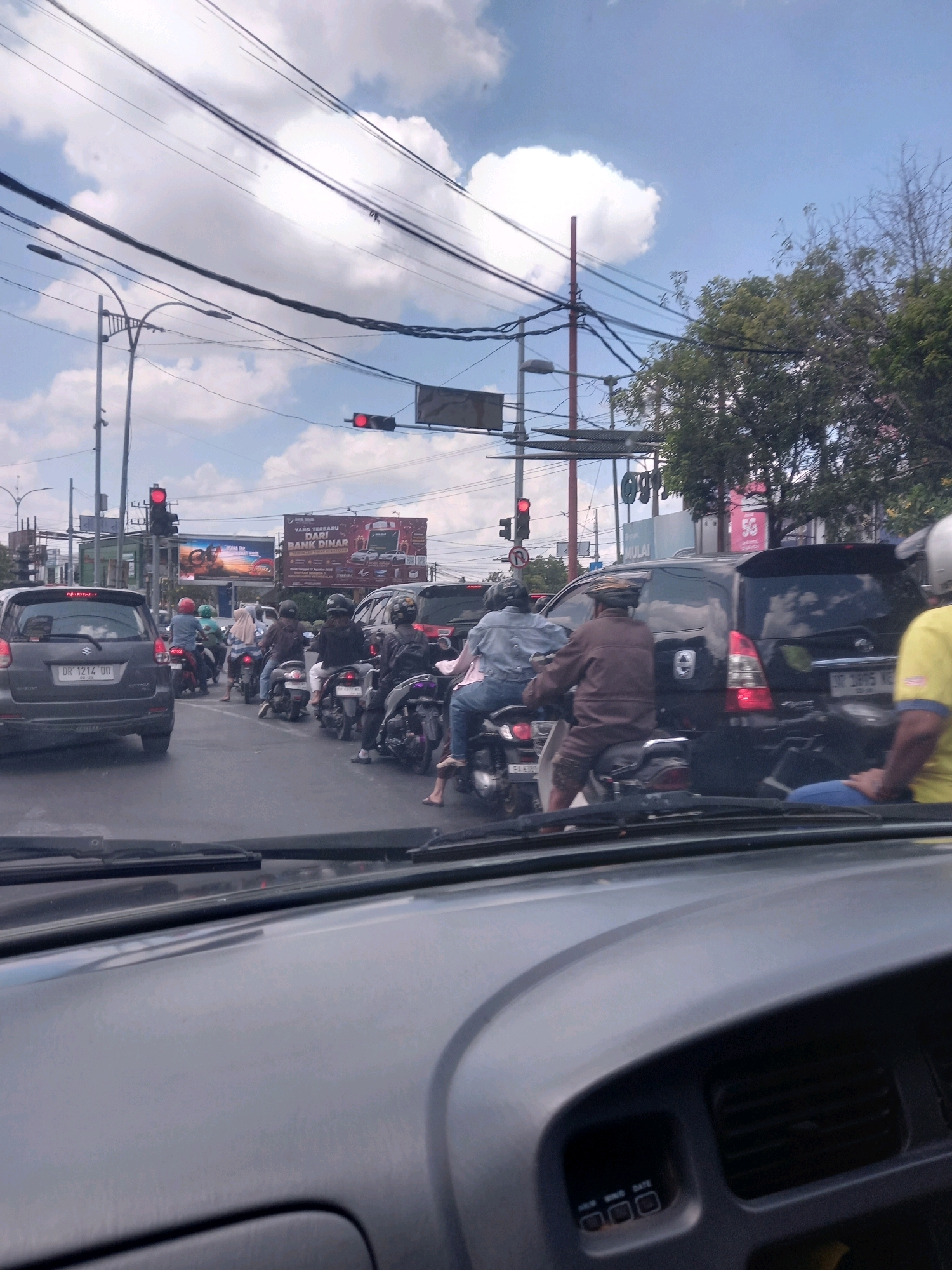 The image depicts a traffic jam on a sunny day in Mataram, Indonesia. A variety of vehicles, including cars and motorcycles, are stopped on a paved road. The foreground shows the interior of a car, including its dashboard and windshield wipers. In the background, there are traffic lights, overhead power lines, streetlights, trees, and buildings. Several people are visible riding on the motorcycles, wearing helmets. One person is wearing a yellow shirt. The sky is blue with scattered white clouds. One traffic light in the image is red.