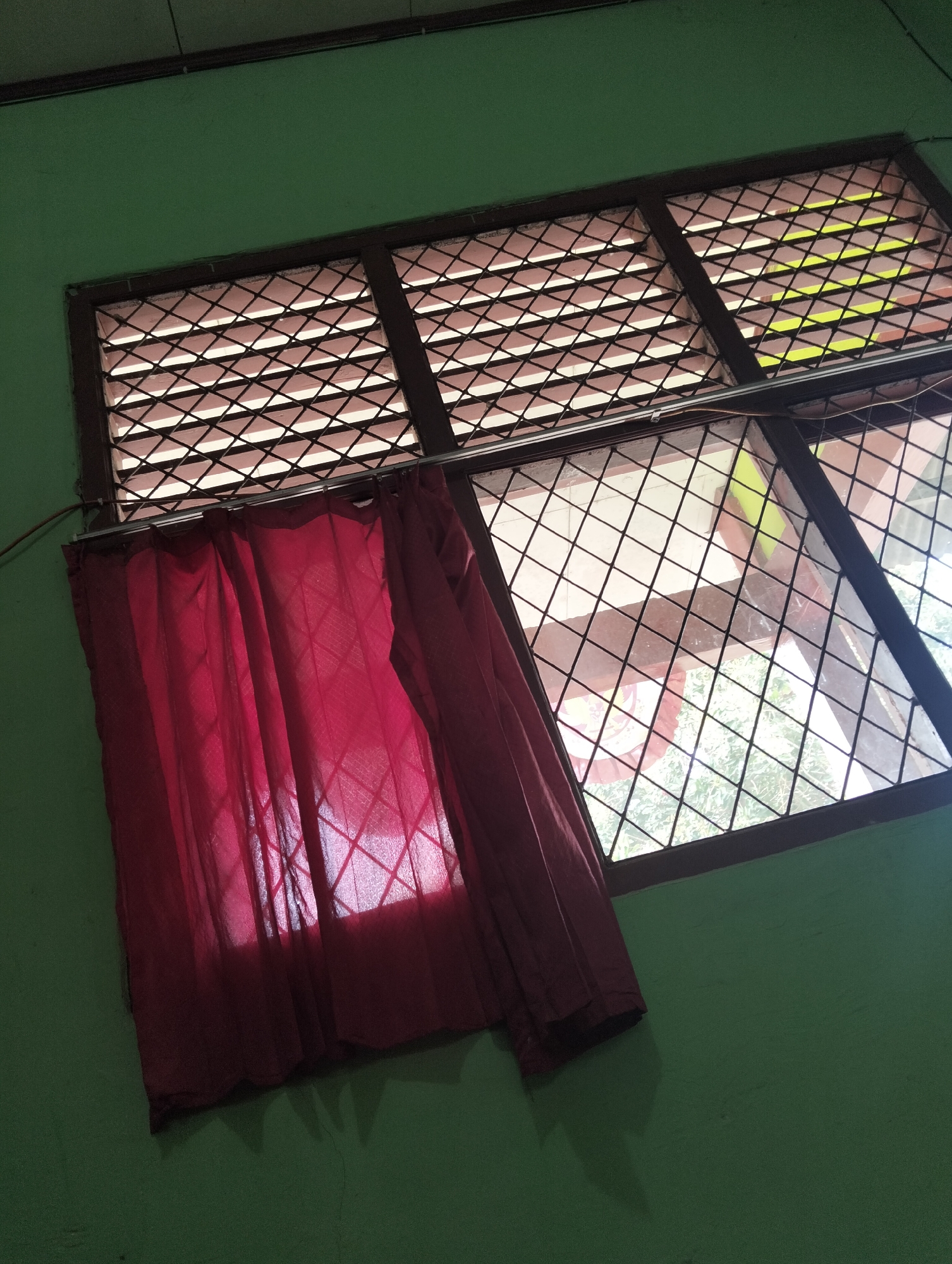 A tilted medium shot captures a window with a lattice and a red curtain against a green wall. The window is divided into three sections horizontally and appears to have bars or a diamond-shaped lattice. The red curtain is partially drawn, covering the left side of the window, and is illuminated by light filtering through. Outside the window, greenery and part of a building are visible. The green wall shows signs of wear and tear. The scene is likely indoors, possibly in a residential or communal setting in Banjar Agung, Indonesia.