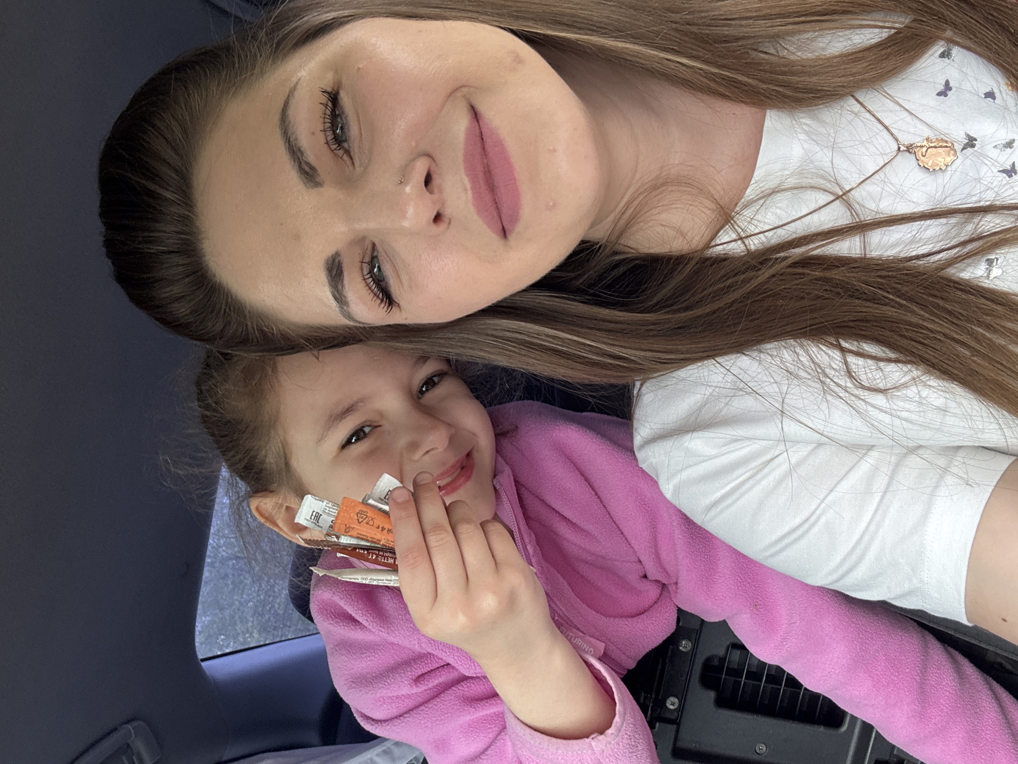 A selfie shows a woman and a child. The woman has long, light brown hair and is wearing a white t-shirt and gold necklace. The child is wearing a pink fleece jacket and holding several small packets. They are inside a car. The location, Shevlyakovo, Russia, cannot be confirmed from the image.