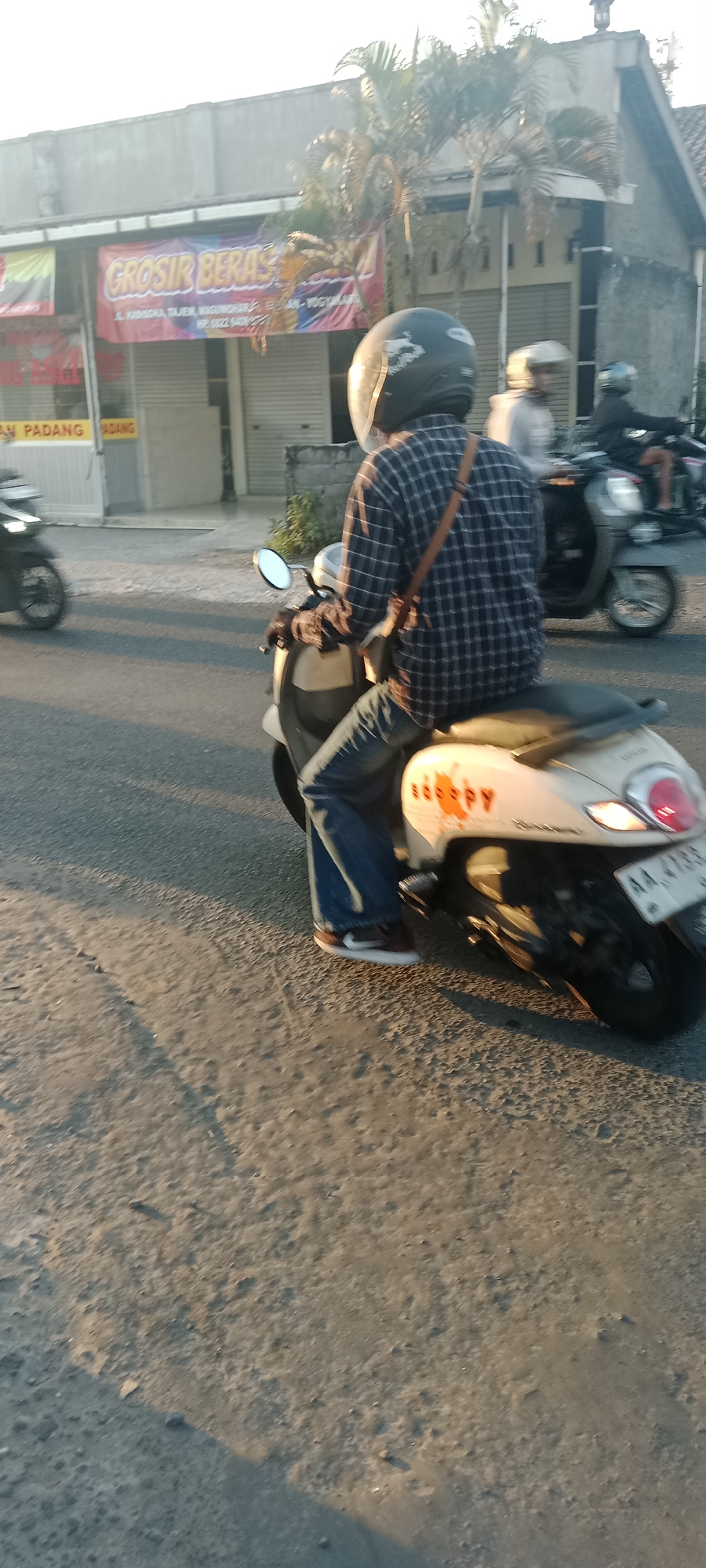 A person wearing a plaid shirt and jeans rides a scooter in Maguwoharjo, Indonesia. The scooter is white with orange decals that read "HAPPY." The person is wearing a black helmet with a reflective visor. In the background, a building with a sign reading "GROSIR BERAS" (Rice Wholesaler) is visible, along with other shops and palm trees. Another scooter is seen in the distance. The scene appears to be a street with asphalt pavement, and the lighting suggests it is late afternoon or early evening.