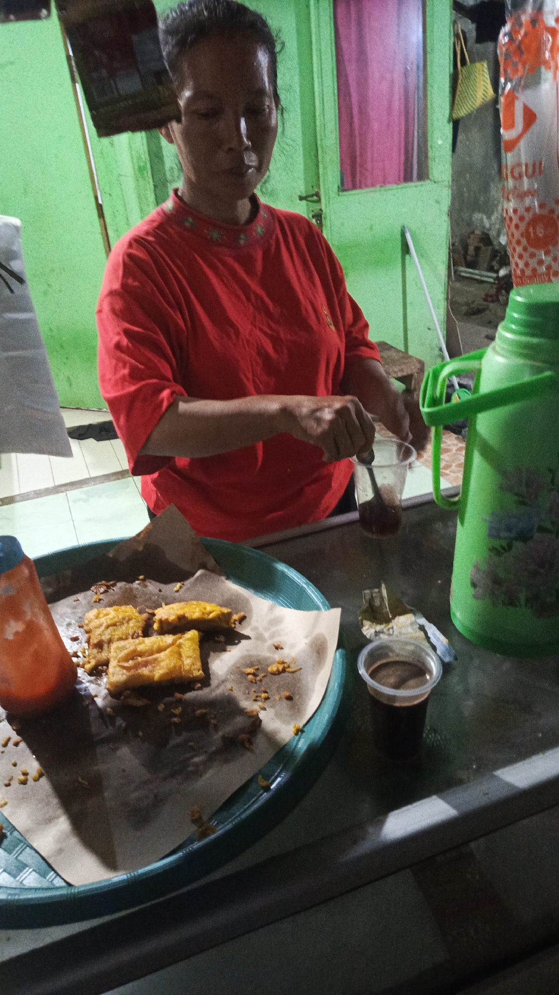 A woman in a red t-shirt is standing behind a table in what appears to be a street food stall. She is in the process of stirring a dark liquid in a clear plastic cup with a spoon. In front of her on the table is a green tray filled with pieces of fried food, possibly tempeh or banana fritters, served on brown paper. To the right of the tray, there is another small plastic cup containing a dark liquid, likely coffee or tea. A large green thermos is also visible on the right side of the image, along with a plastic bottle that appears to be a ketchup dispenser. The background shows a green painted wall and a doorway with pink curtains. The lighting suggests it is either dusk or an indoor setting with artificial light. The overall scene depicts a food vendor preparing or serving snacks and beverages, likely in a casual outdoor or semi-outdoor setting in Tangerang, Indonesia.