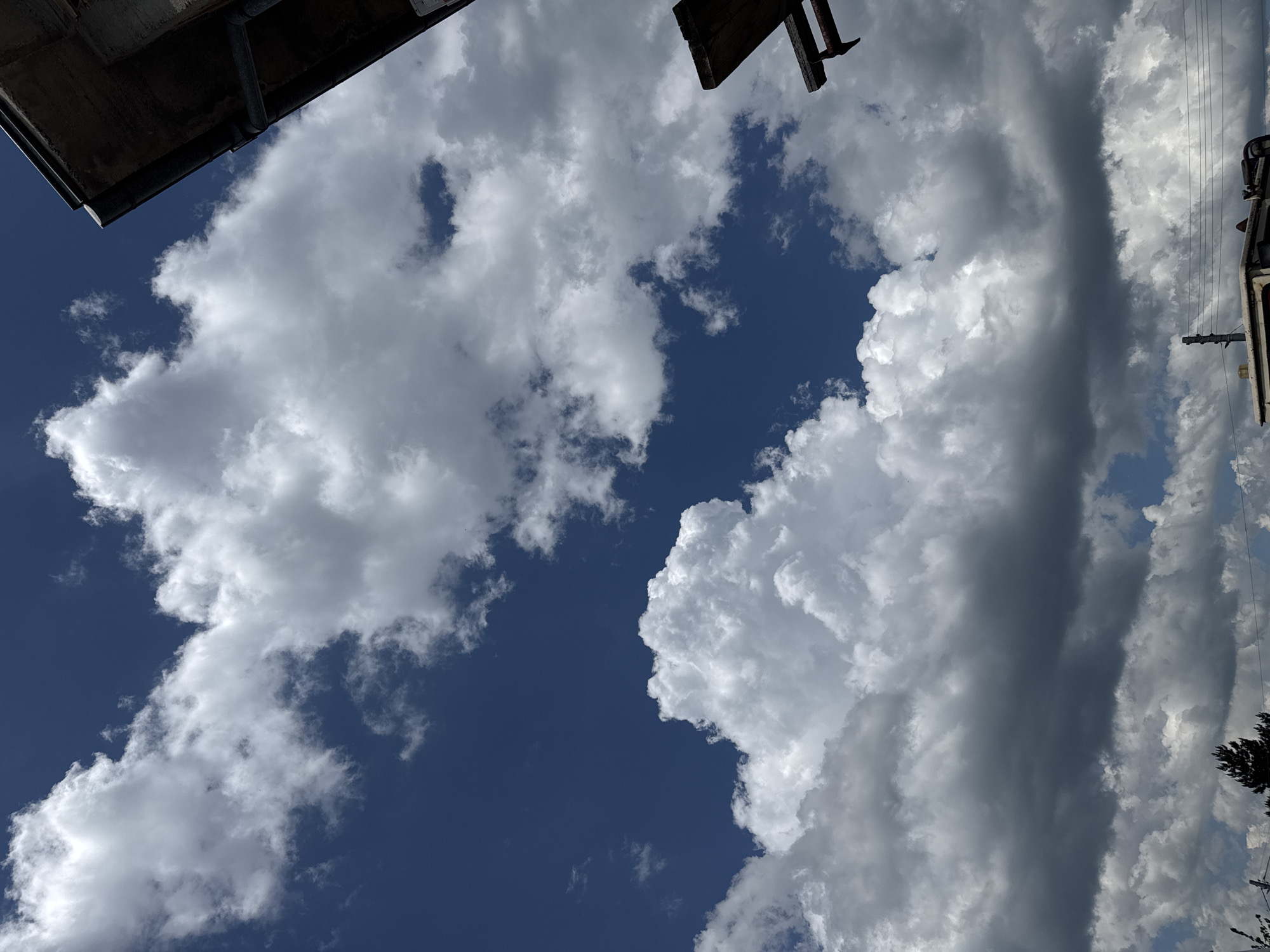 The image shows a low-angle view of the sky above Sarnevo, Bulgaria. Puffy white cumulus clouds are scattered across a deep blue sky. In the upper left and right corners, parts of buildings with dark, possibly metal, structures are visible. On the right side, electrical wires and a utility pole are visible against a background of lighter, thinner clouds. Some trees can be seen at the bottom right.