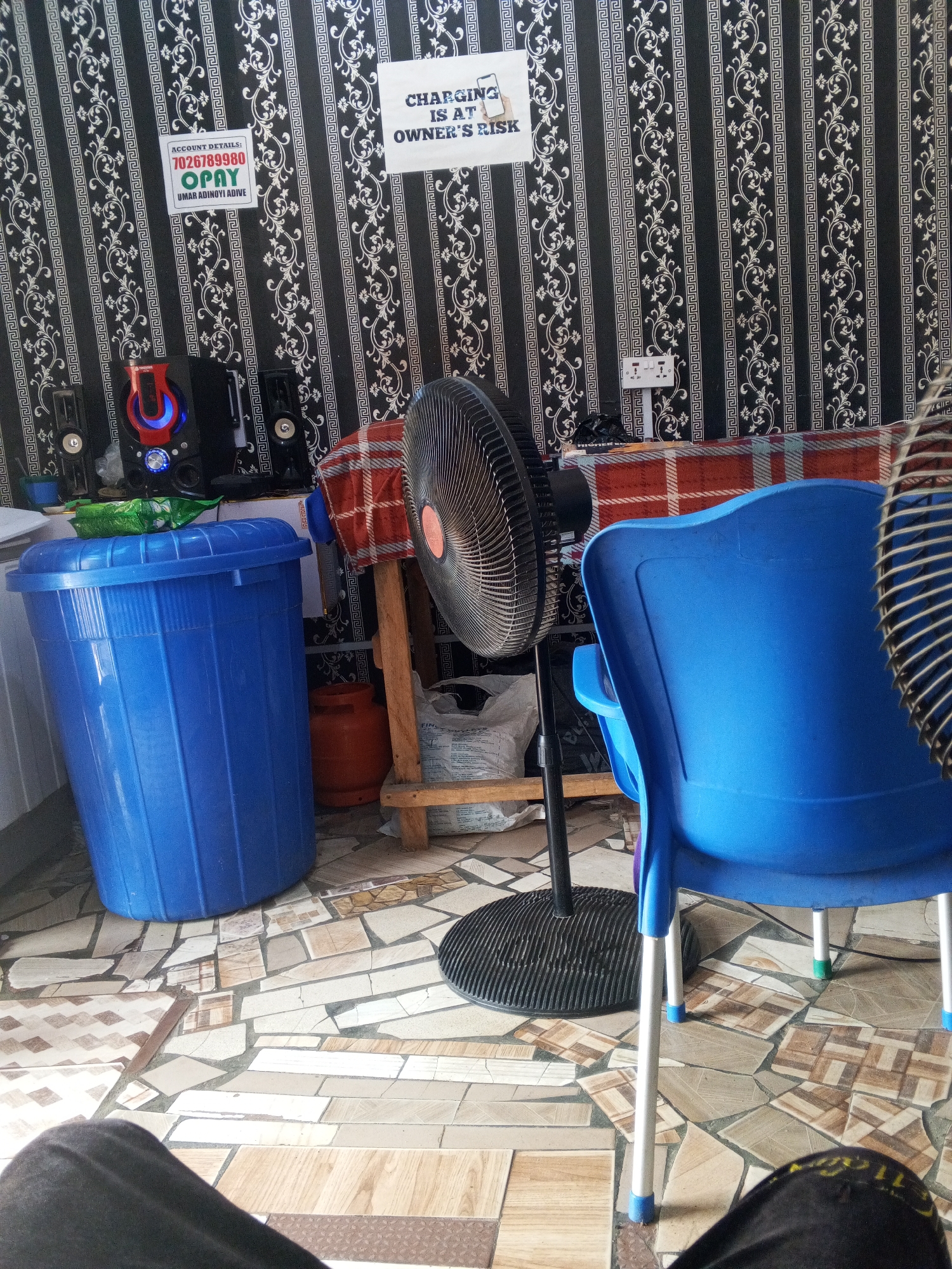 The interior of a room in Lokoja, Nigeria, presents a cluttered scene. The walls are covered with patterned wallpaper, and a sign that reads "CHARGING IS AT OWNER'S RISK" is displayed alongside a notice with the account details "7026789980 OPAY UMAR ADINOYI ADIVE." A computer speaker system is placed on a surface draped with a red and orange patterned cloth. To the left, a large blue plastic trash can stands next to a small orange propane tank. In the foreground, a black standing fan and a blue plastic chair dominate the view. The patterned tile floor is visible between the objects. A person's lap, clad in dark trousers, is seen in the bottom foreground.