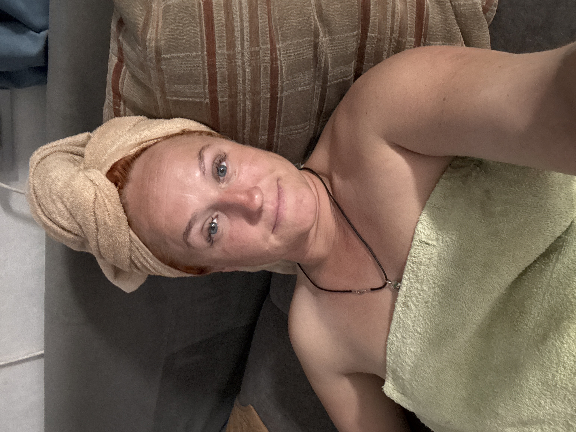 A red-haired woman with blue eyes is lying on a couch, wrapped in towels. She has a towel turban on her head and another towel wrapped around her body. A necklace with a pendant is visible around her neck. The couch appears to be in a domestic setting, possibly a living room. There is a pillow with a striped pattern next to her. The lighting suggests it is evening or indoors. The specific location in the city of Chaikovsky, Russia cannot be determined from the image alone.