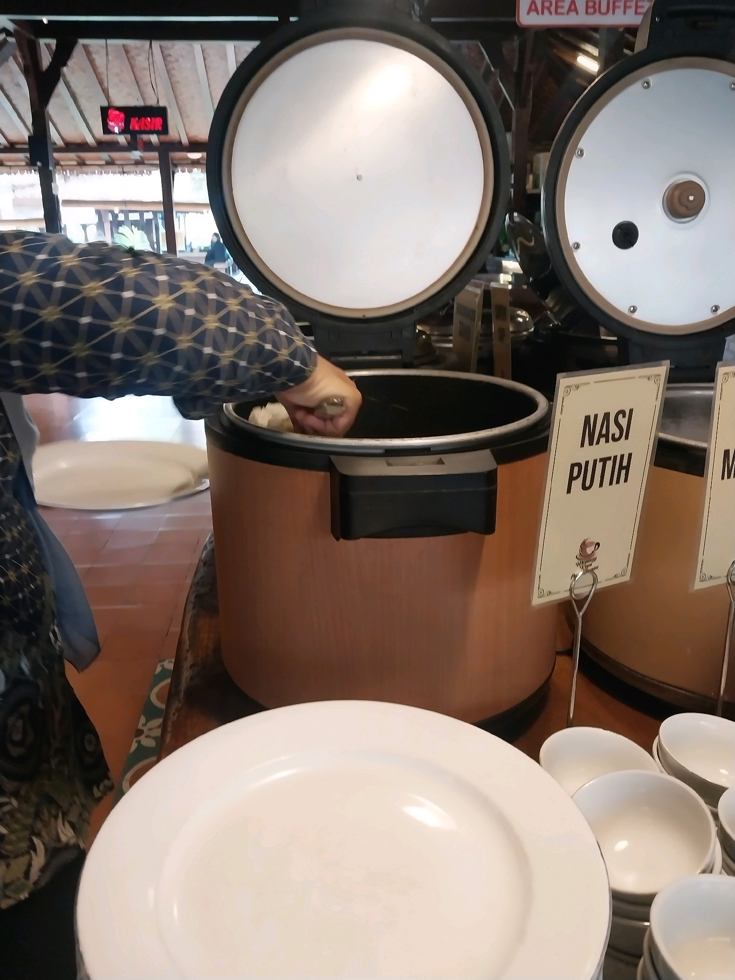 This image depicts a buffet serving area, likely in Bugisan, Indonesia. In the foreground, a stack of white plates and bowls are visible on a wooden surface. Behind these, a person in a dark, patterned long-sleeved shirt is using a ladle to serve food from a large, brown, cylindrical container with an open lid. This container appears to be a rice cooker or a food warmer. A white sign with black text reading "NASI PUTIH" (white rice in Indonesian) is displayed in front of a similar container to the right. The background shows more buffet equipment and a ceiling structure. The overall scene suggests a self-service dining environment.