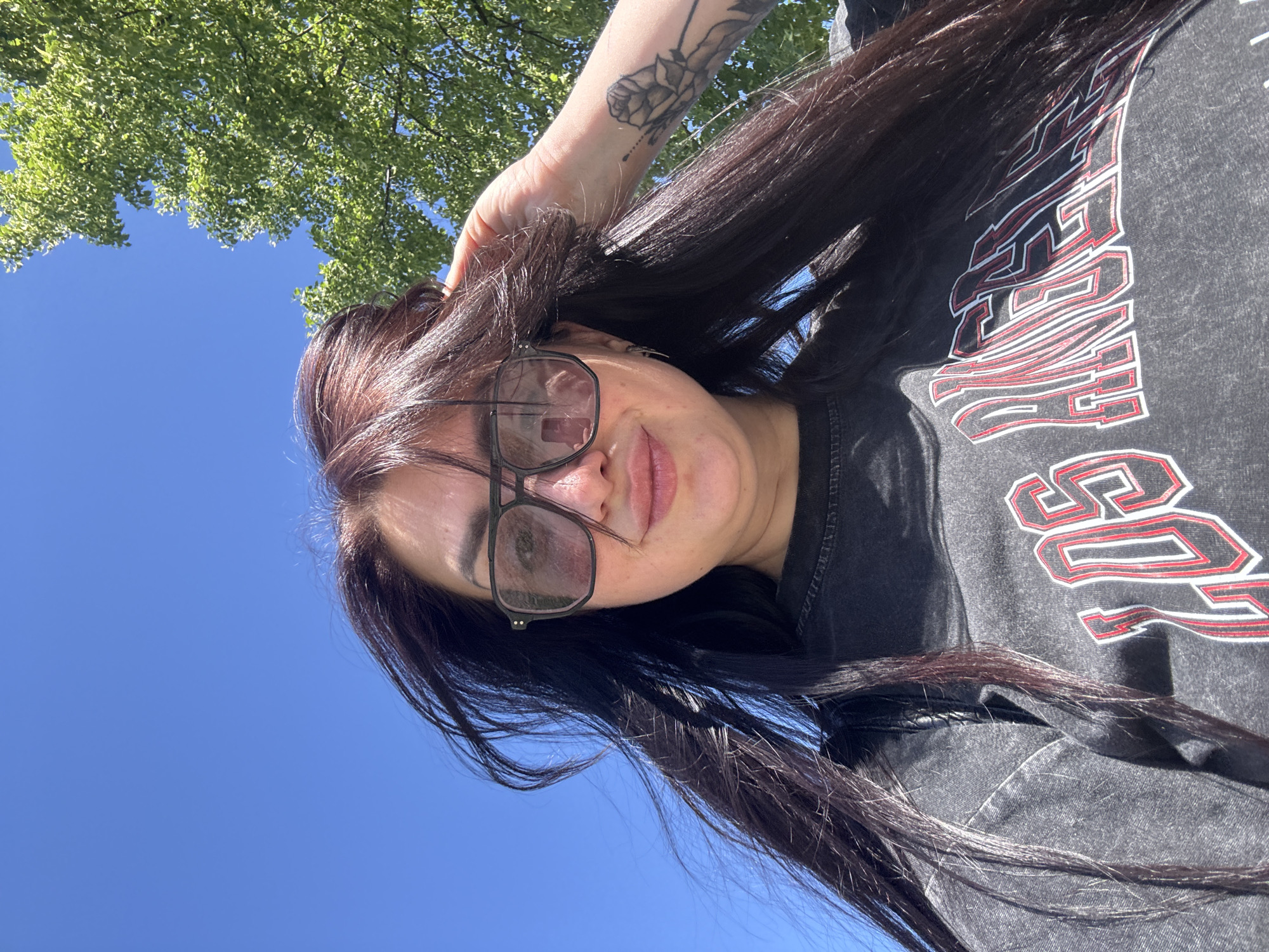 A low-angle medium shot shows a woman with long, dark hair and eyeglasses standing outdoors in Unknown, Unknown. The woman is wearing a black t-shirt with white and red text, and her left arm is raised towards her head, with a tattoo visible on her forearm. Behind the woman is a clear blue sky and the foliage of a tree.