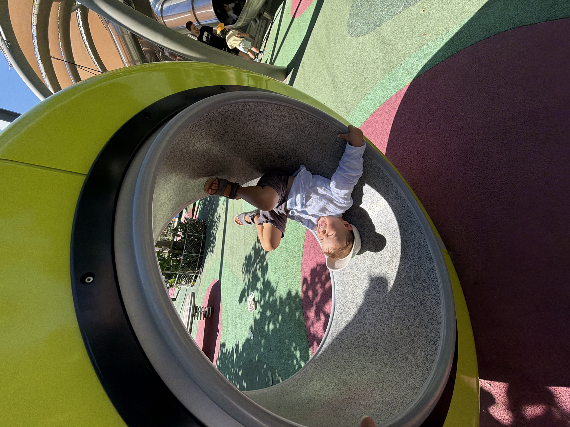 A young child lies upside down inside a curved, cylindrical tunnel at a playground. The tunnel is made of grey, speckled material with a lime green outer shell. The child is wearing a white long-sleeved shirt, dark shorts, sandals, and a white hat. Their legs are bent, and their arms are resting on the edge of the tunnel. The playground has a rubberized surface in shades of pink and green, with shadows from trees cast across it. In the background, other children and playground equipment, including slides and a spring rider, are visible. The scene suggests a sunny day at a public park. Based on the presence of modern playground equipment designed with bright colors and safe, textured surfaces, and the style of the surrounding architecture, this playground is likely located in a contemporary urban park within Barcelona, Spain, known for its family-friendly amenities.