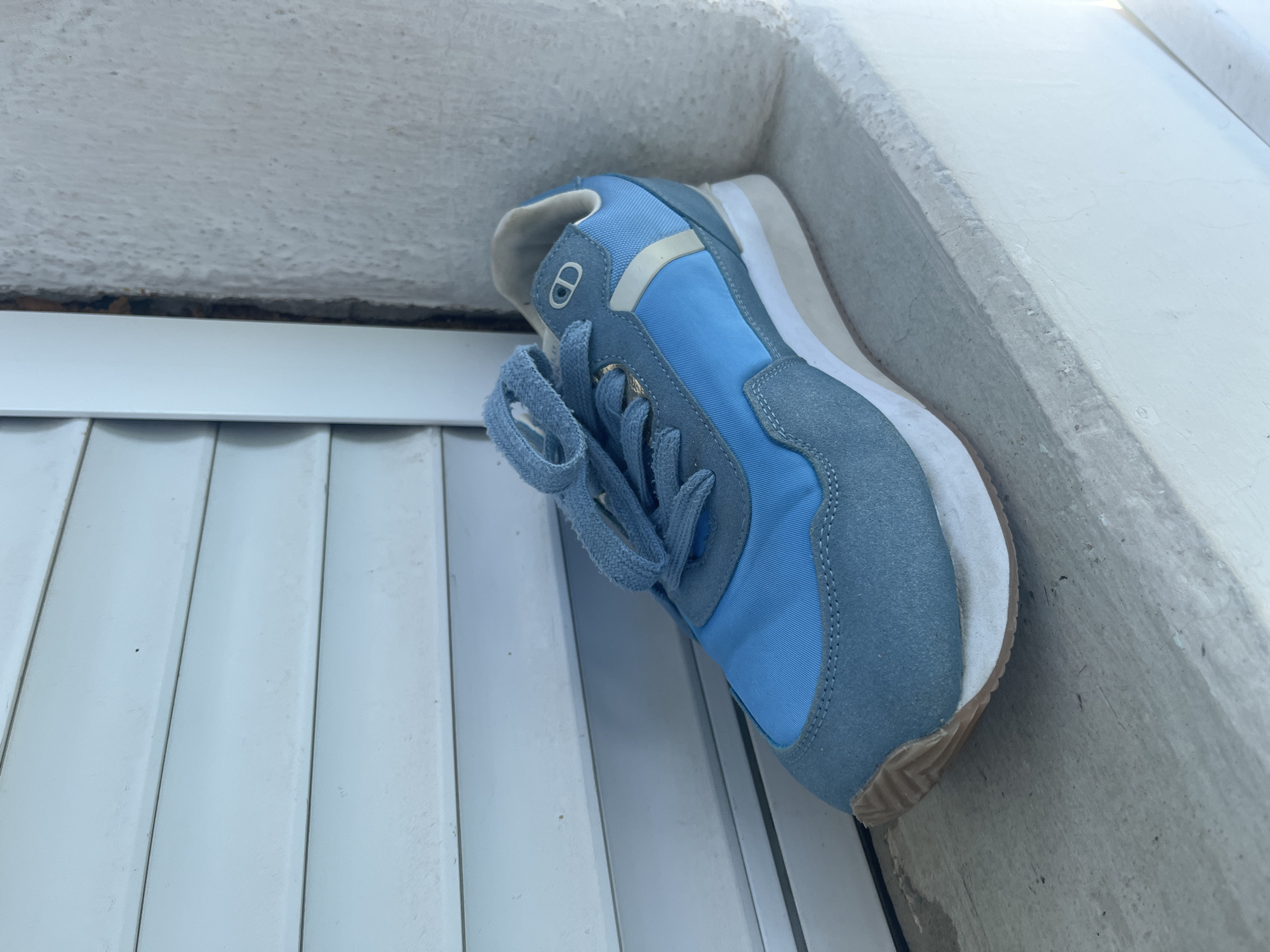 A light blue athletic shoe rests on a white, textured ledge. The shoe is positioned vertically, with its sole facing down and its laces loosely tied. The shoe has a lighter blue textile on the toe box and side panels, and a darker blue suede on the heel and eyelet area. A light beige stripe runs across the side of the shoe. The sole of the shoe is white with a tan rubber outsole.
The shoe is placed against a rough, white, stucco-like surface that forms a corner or ledge. Below and to the left of the shoe, a series of parallel white slats or panels are visible, suggesting an outdoor setting, possibly a patio or balcony. The lighting suggests it is daytime, with natural light illuminating the scene. The background is out of focus but appears to be an outdoor environment. There is no text visible in the image. The overall impression is one of a casual, perhaps forgotten, object in an outdoor space.