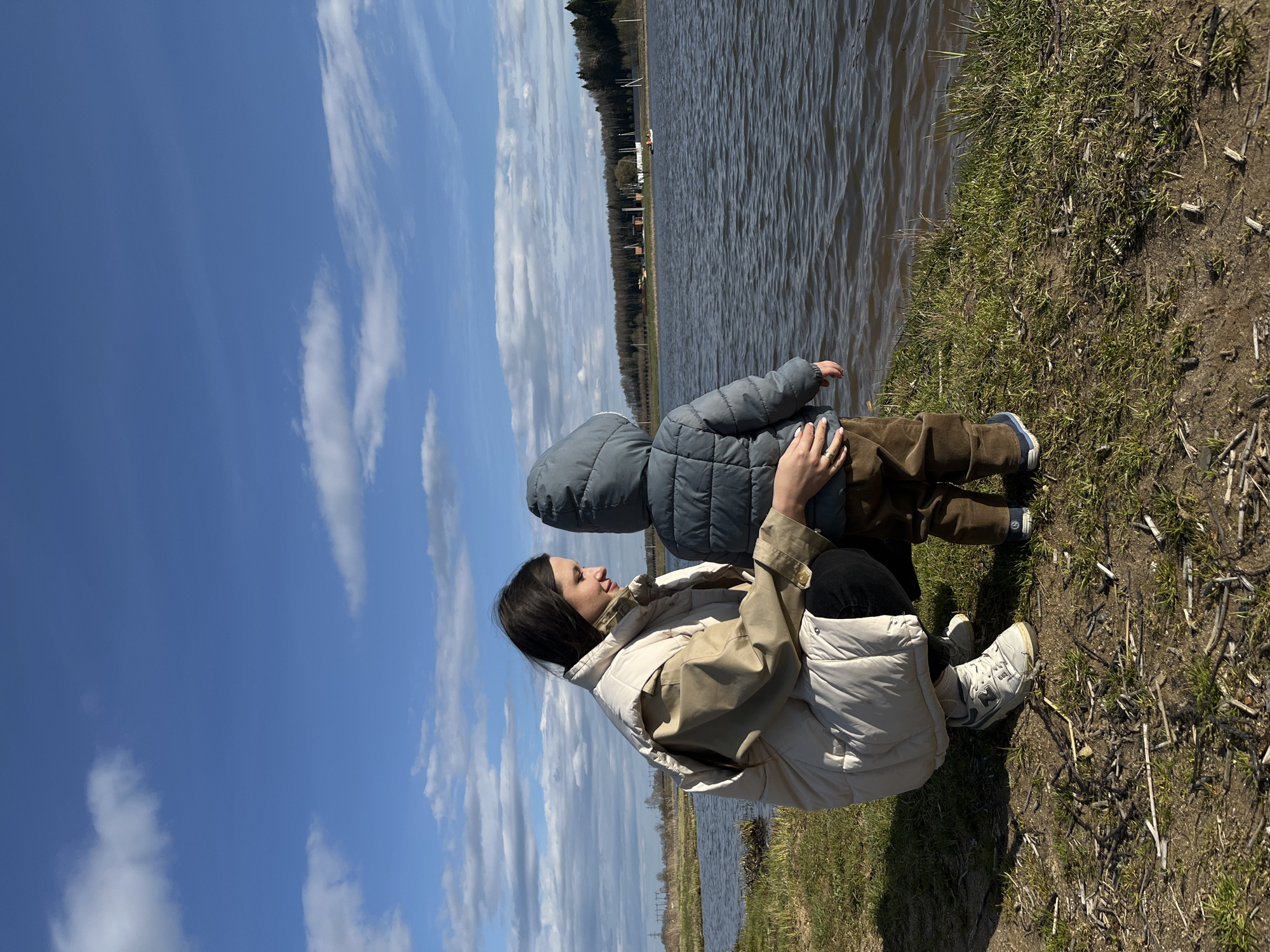 In Литвиново, Russia, a woman in a beige and white jacket is kneeling next to a child wearing a blue hooded jacket and brown pants. They are positioned on the bank of a body of water, possibly a river or lake. The shoreline has grassy vegetation and dry stems. Beyond the water, a line of trees is visible in the distance. The sky is blue with scattered white clouds. The woman appears to have dark hair and is looking towards the water. Her hand is placed on the child's back.