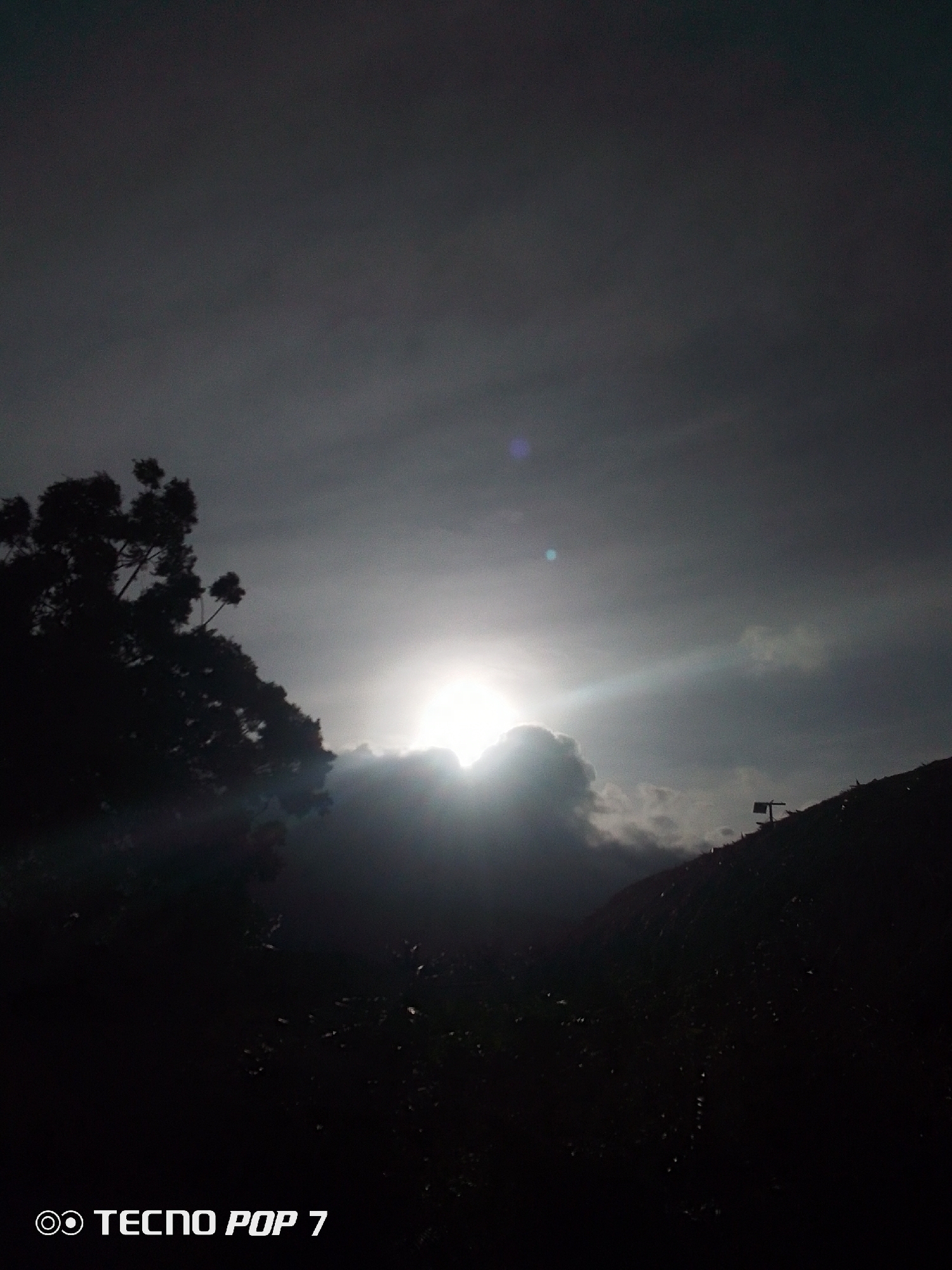 The image depicts a landscape near Pedro González, Venezuela, under a dark, overcast sky. The sun, obscured by clouds, shines brightly through a gap, creating a bright spot in the center of the frame and rays of light across the sky. To the left, the silhouette of a tree with sparse foliage is visible. The foreground is dominated by dark, dense foliage, with a hillside visible on the right side of the frame. A structure, possibly a small shed or pole, is positioned on the hillside. The environment is predominantly dark and shadowy, suggesting either a pre-dawn or dusk setting, or a day with heavy cloud cover. At the bottom of the frame, the text "TECNO POP 7" is visible, indicating the device used to capture the image.