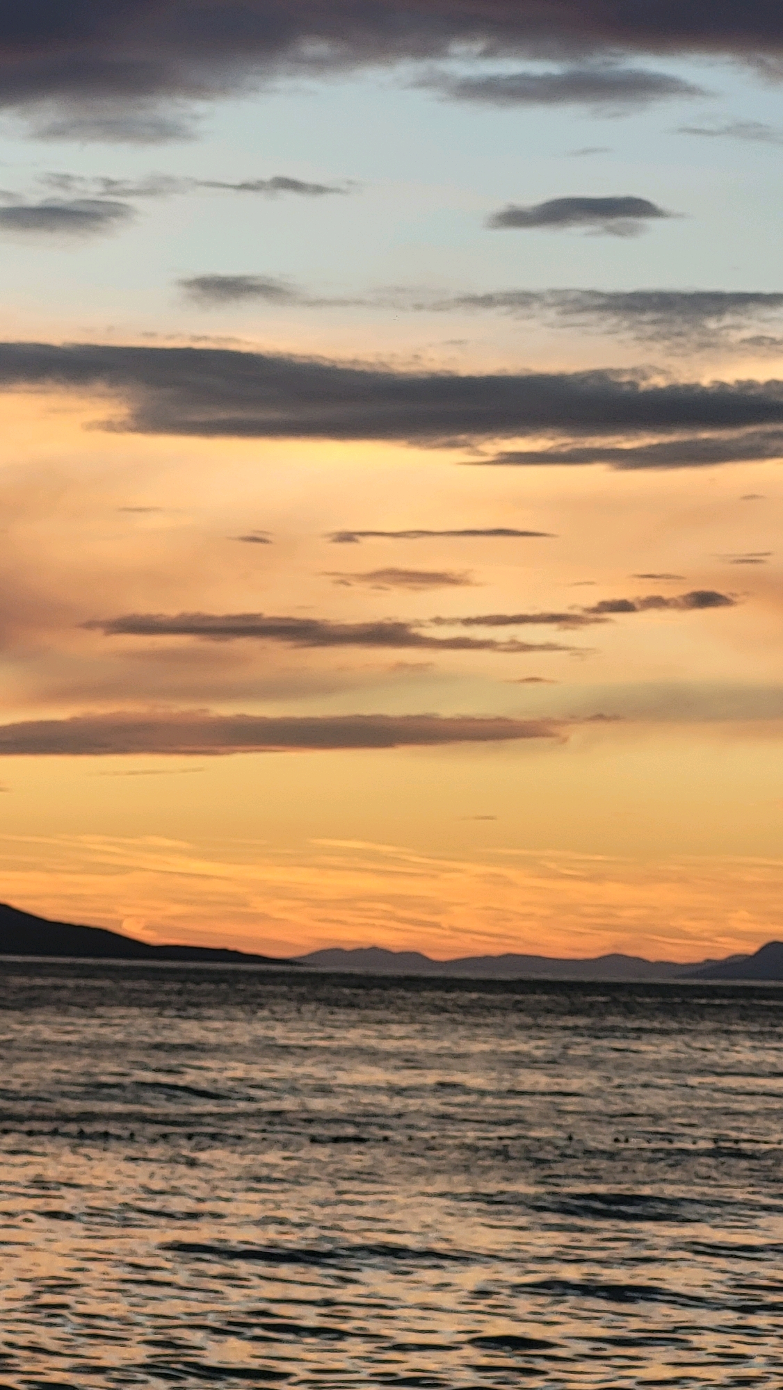 The image depicts a serene seascape at dusk, with the sky painted in warm hues of orange, yellow, and hints of purple and grey. Wispy clouds streak across the horizon, some dark and others light, creating a dramatic contrast against the fading sunlight. Below the sky, a dark silhouette of distant mountains forms the backdrop. In the foreground, a calm body of water, likely the Adriatic Sea, reflects the colors of the sky, with gentle ripples disturbing the surface. The overall impression is one of tranquility and natural beauty. The location is not specified within the image itself.