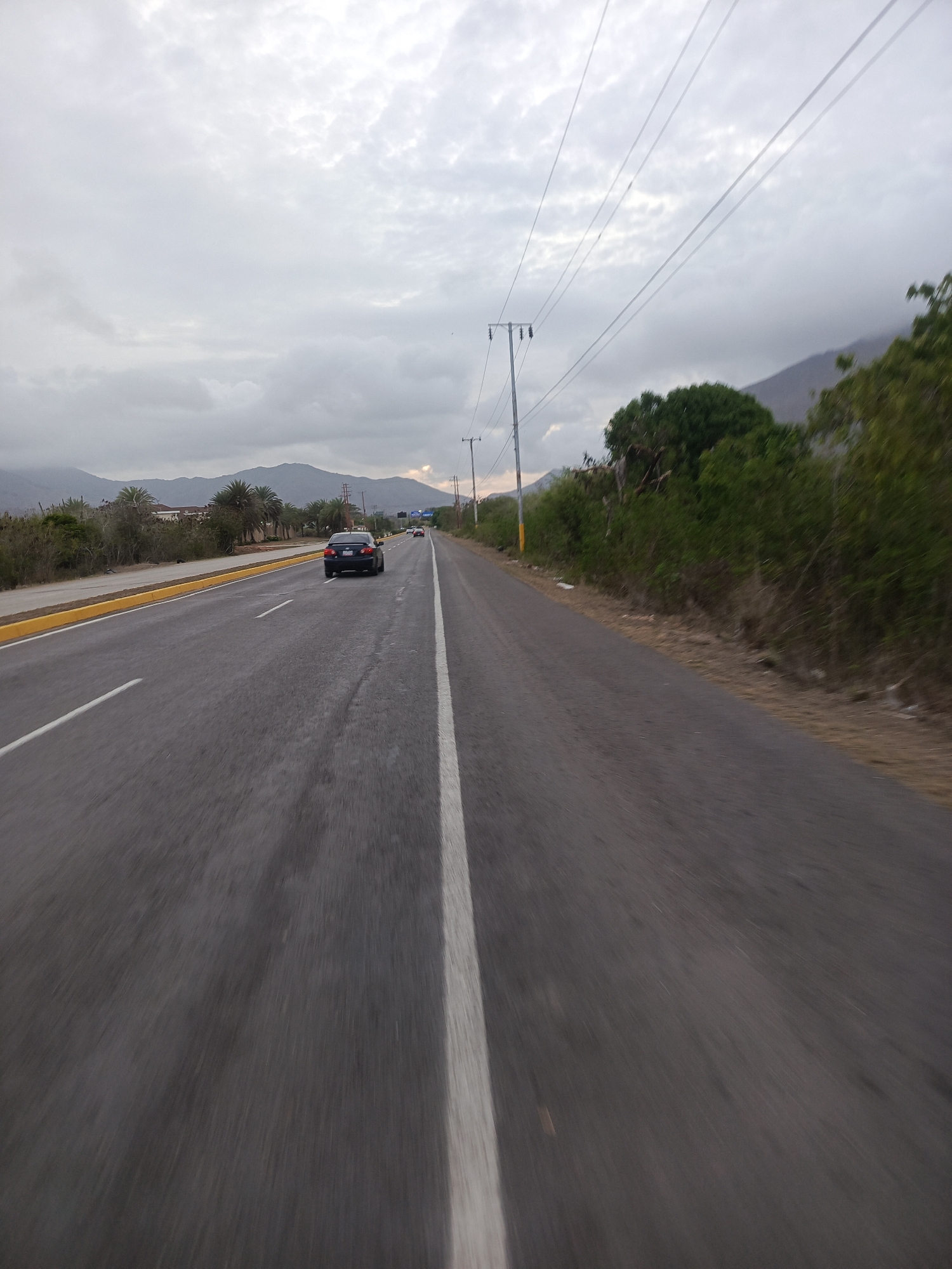 This image captures a wide-angle view of a multi-lane asphalt highway in La Asuncion, Venezuela, under an overcast sky. The road features a visible lane in the foreground with a solid white edge line, leading to a central median defined by yellow curbs and sparse green vegetation, including palm trees. Beyond the median, another road segment is visible, bordered by dry scrub, additional greenery, and indistinct distant structures.
A black sedan is observed in the distant left lane, moving away from the viewer, with a lighter-colored vehicle further ahead. To the right of the foreground lane, a dirt shoulder exhibits dry vegetation and minor debris, succeeded by a dense line of green bushes and trees. Several metal utility poles, carrying multiple overhead power lines, run parallel to the road on the right side. The background is dominated by a range of hazy, grey-blue mountains stretching across the horizon. The sky is heavily clouded, presenting diffused, subdued lighting, with a faint orange glow near the horizon on the far right, possibly indicating a hidden sunrise or sunset. No people are visible outside of the vehicles.