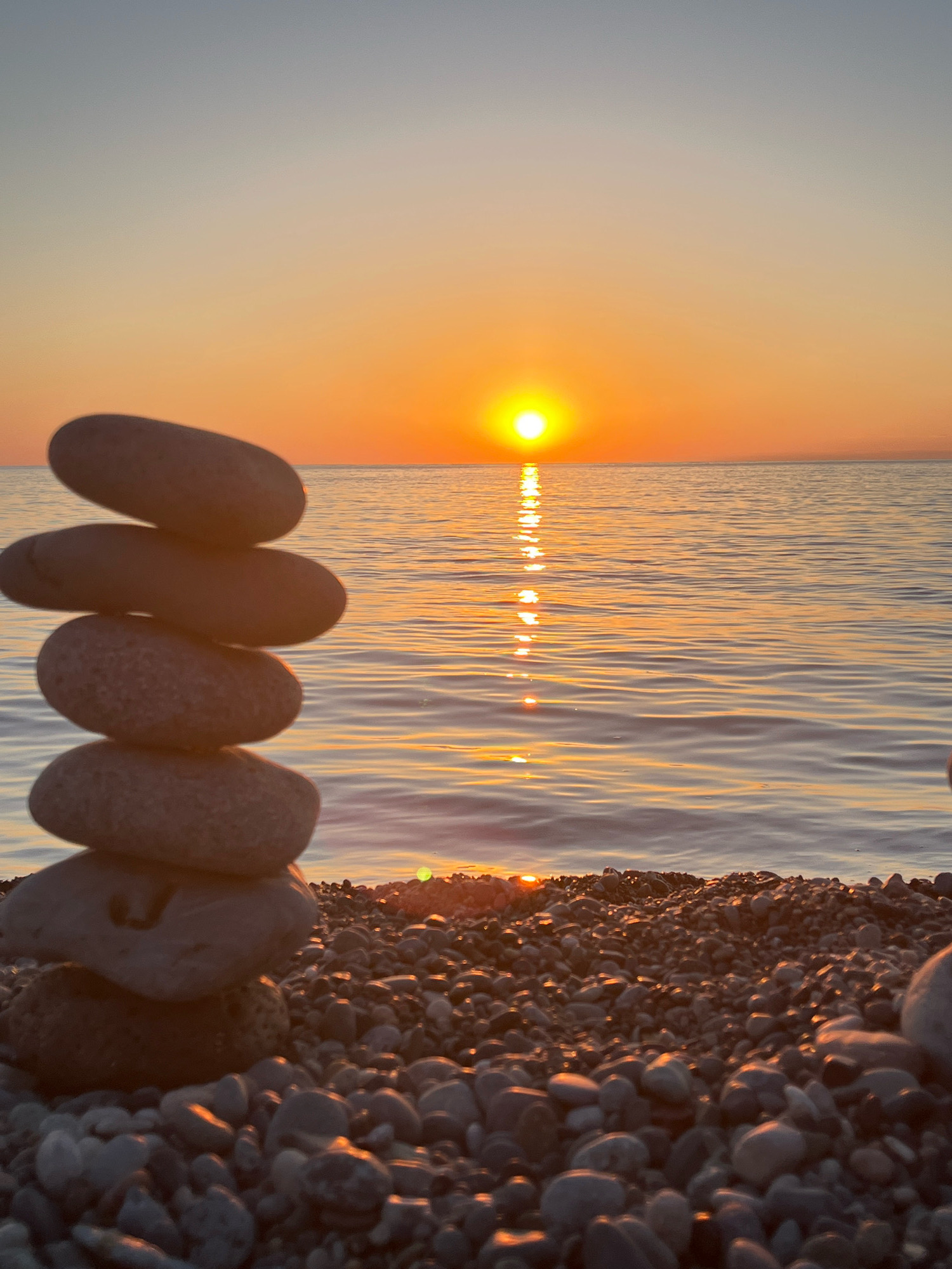 The image captures a serene sunset scene on a pebble beach in Kobuleti, Georgia. A stack of five smooth, gray stones is balanced in the foreground, with the largest stone at the bottom and progressively smaller stones stacked on top. Beyond the stones, the calm sea stretches towards the horizon, reflecting the warm, orange glow of the setting sun. The sun, a bright orb, is positioned slightly to the left of the center of the frame, casting a shimmering path of light across the water. The sky transitions from a soft blue at the top to a vibrant orange and yellow near the horizon, creating a peaceful and tranquil atmosphere. The beach itself is covered in a multitude of small, smooth pebbles of varying shades of gray and brown. The overall impression is one of natural beauty and calm.