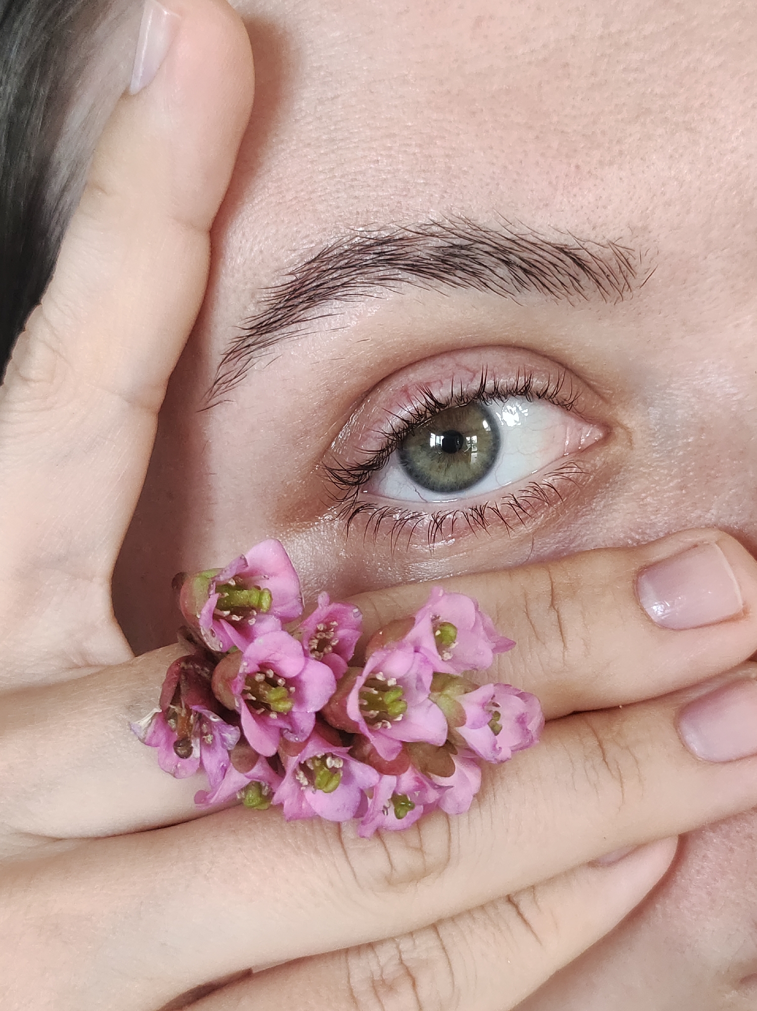A close-up image shows a person's left eye partially obscured by their hand, which is adorned with a cluster of small pink flowers. The eye has green irises, and the skin around the eye is smooth with some visible blood vessels. The eyebrow is dark and well-defined. The flowers, which appear to be a type of bergenia, are positioned between the eye and the fingertips. The fingernails are short and appear to have a light pink or nude polish. The background is out of focus, but suggests the person has dark hair. The location is not discernible as Фоки, Russia based on the provided image.