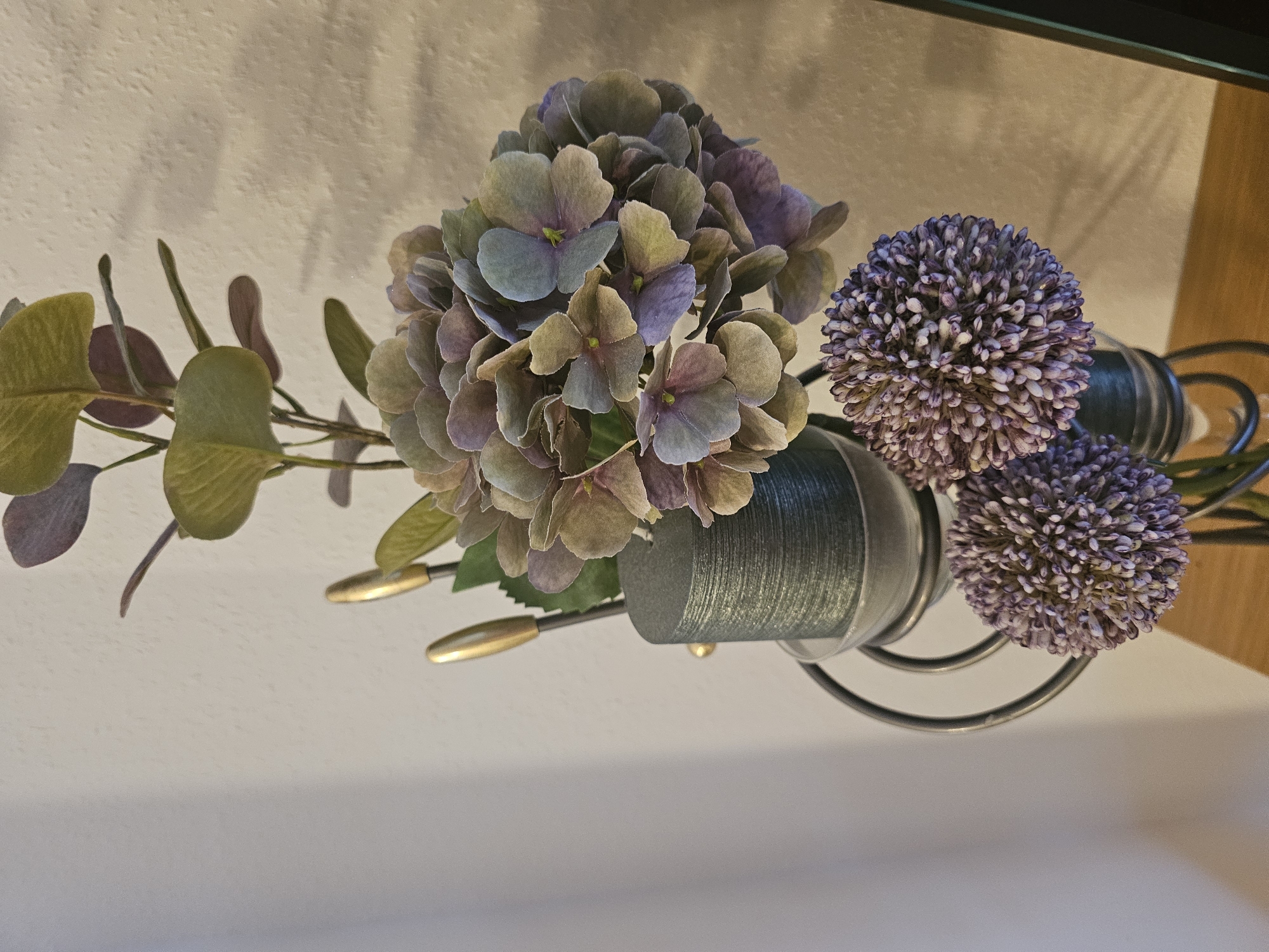 A close-up shot focuses on an artificial floral arrangement and a dark green candle in a metal holder. The arrangement includes a cluster of muted purple and green hydrangea-like flowers and two spherical purple allium-like blooms. Two gold-tipped decorative elements, possibly part of the floral arrangement or the candle holder, are visible behind the hydrangea. The candle is cylindrical and has a textured surface. The arrangement is set against a lightly textured off-white wall, with subtle shadows cast on it. The lower portion of the image shows a wooden surface.