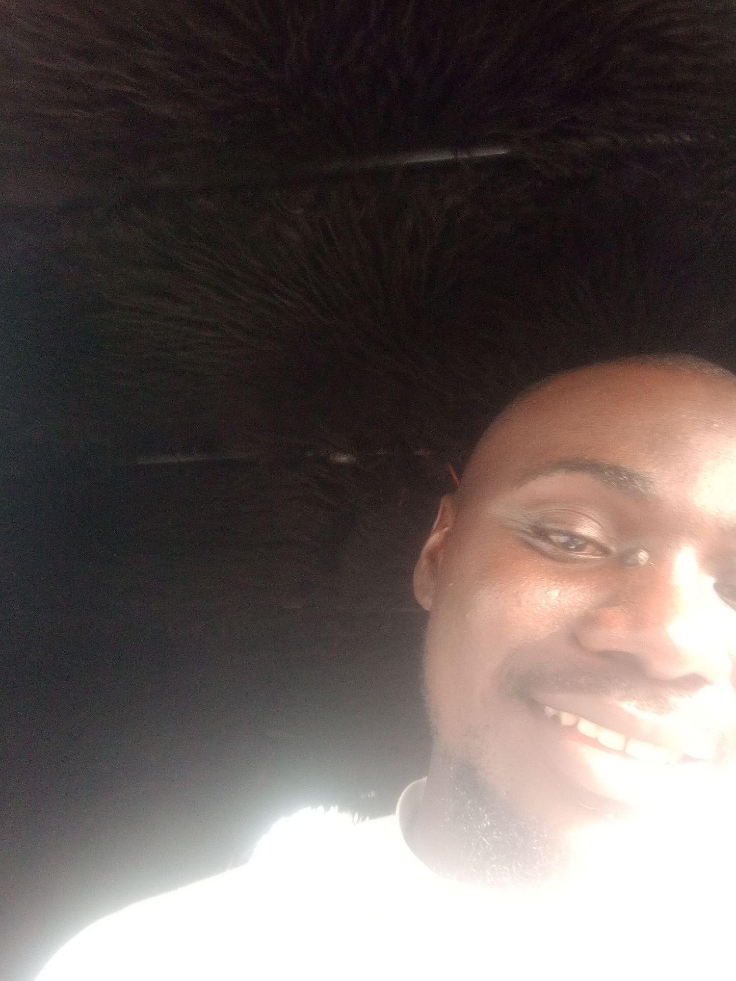 The image is a selfie taken by a Black male with a shaved head and light stubble on his face. He is smiling and looking towards the camera. His right eye is visible and appears to be brown. The lighting in the image is bright, creating a glare that obscures some details of his face. The background is dark and appears to be the interior of a vehicle, possibly with some fuzzy material visible at the top. The image is composed from the chest up. There are no other people or objects clearly visible in the image. It is not possible to determine the location in Jalingo, Nigeria, from the image alone.