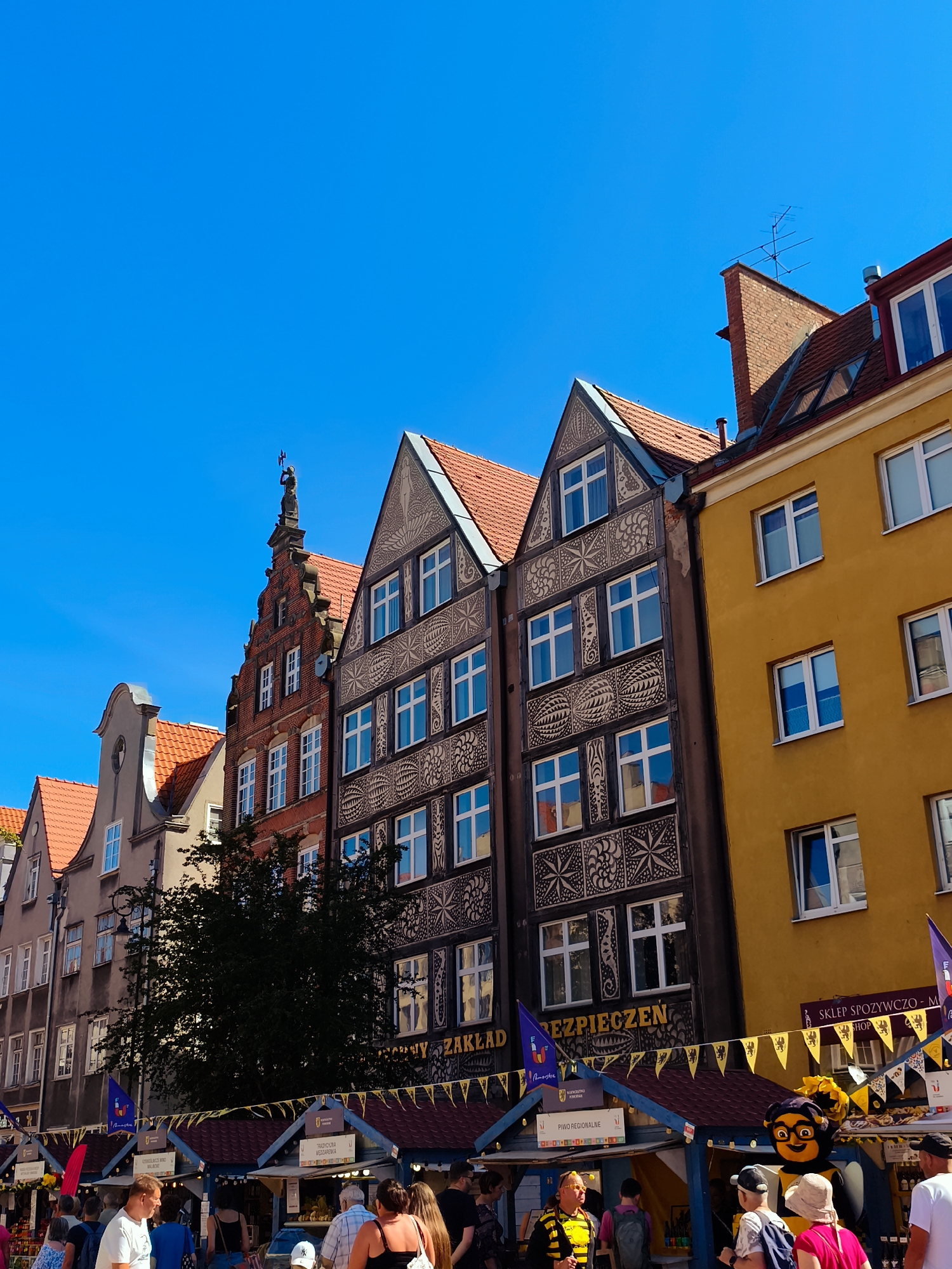 The image captures a street scene in the city of Gdańsk, Poland, likely during a market or festival. The architecture features tall, narrow buildings with red tile roofs and decorative facades. One building stands out with intricate black and white patterns on its upper levels, while another is painted a vibrant yellow. In the foreground, there are market stalls with red roofs, adorned with yellow pennant banners. People are visible browsing the stalls and walking along the street, dressed casually in summer clothing. The sky is a clear, bright blue. The overall impression is of a lively, bustling urban environment on a sunny day.