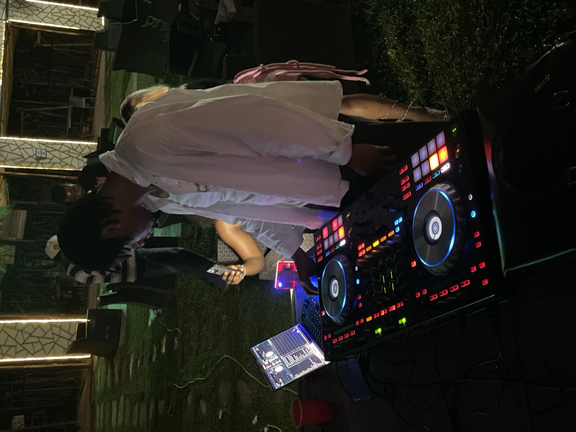 A person is operating a DJ console at night in an outdoor, grassy area. The DJ, wearing a white shirt, is positioned in the foreground, with their hands on the console. A laptop displaying DJ software is open to the left of the console. In the background, other individuals are visible, including one person holding a smartphone. The environment appears to be a venue or gathering space, possibly in Abuja, Nigeria, with illuminated architectural elements and seating areas. The lighting suggests it is evening or night.