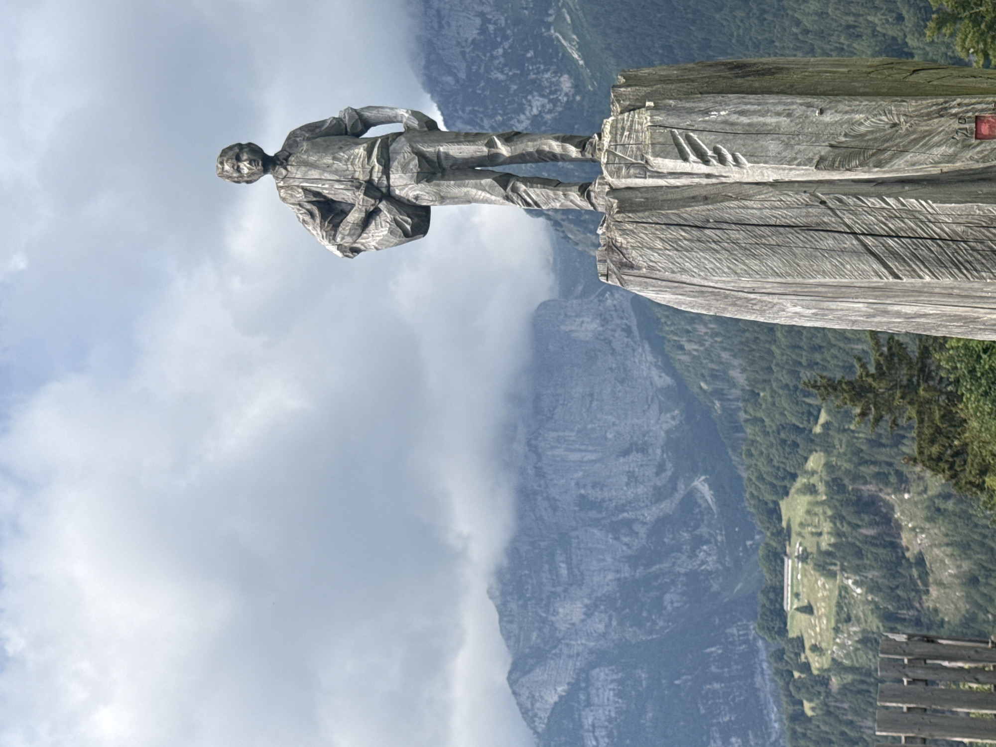 A wooden statue of a young boy carrying a sack over his shoulder stands on a wooden pillar in the foreground. The statue is carved from a single piece of wood, with visible grain and tool marks. The boy is dressed in a shirt and trousers, and he appears to be looking out at the landscape. The wooden pillar is also carved with intricate patterns, suggesting it is part of the artwork.
In the background, a majestic mountain range rises, its peaks partially obscured by clouds. The mountains are covered in dense green forest, with a few patches of bare rock visible. A small building with a white roof is situated on a grassy hillside amongst the trees, suggesting a rural or alpine setting. A wooden fence runs along the bottom left of the frame, partially obscuring the view of the landscape. The sky above is a mixture of blue sky and dramatic, cloudy formations.
This statue is located in Molveno, Italy, likely on a hiking trail or viewpoint, offering panoramic views of the surrounding Dolomite mountains. The scene captures a sense of natural beauty and traditional craftsmanship.