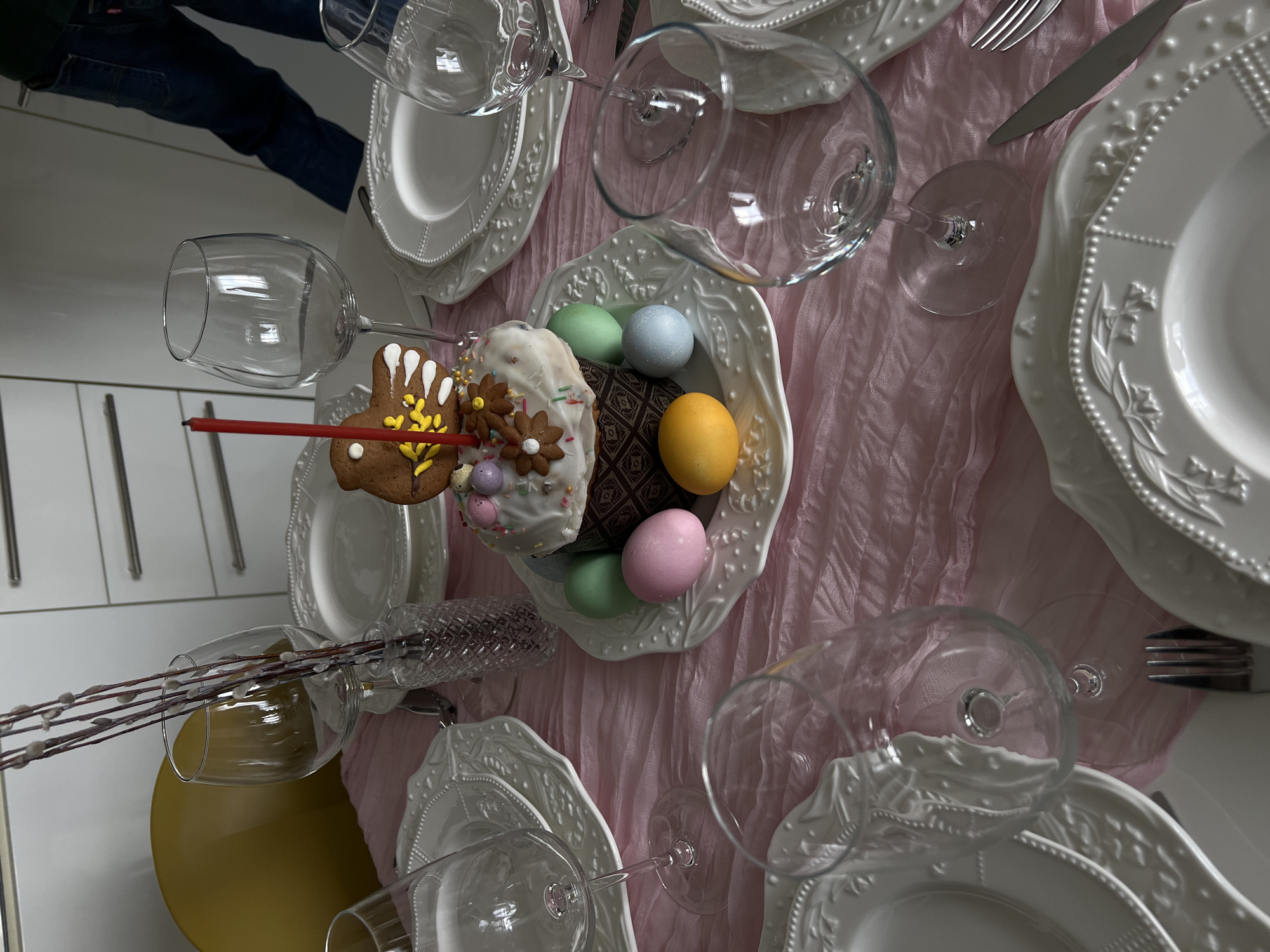 The image depicts a table setting, possibly for Easter, with white decorative plates, wine glasses, silverware, and a light pink tablecloth. In the center of the table, there is a decorative plate containing an Easter cake, a gingerbread bunny with a red candle, and several colored eggs. The setting is indoors, likely a dining room. It is not possible to determine the location as Khimki, Russia from the image alone.