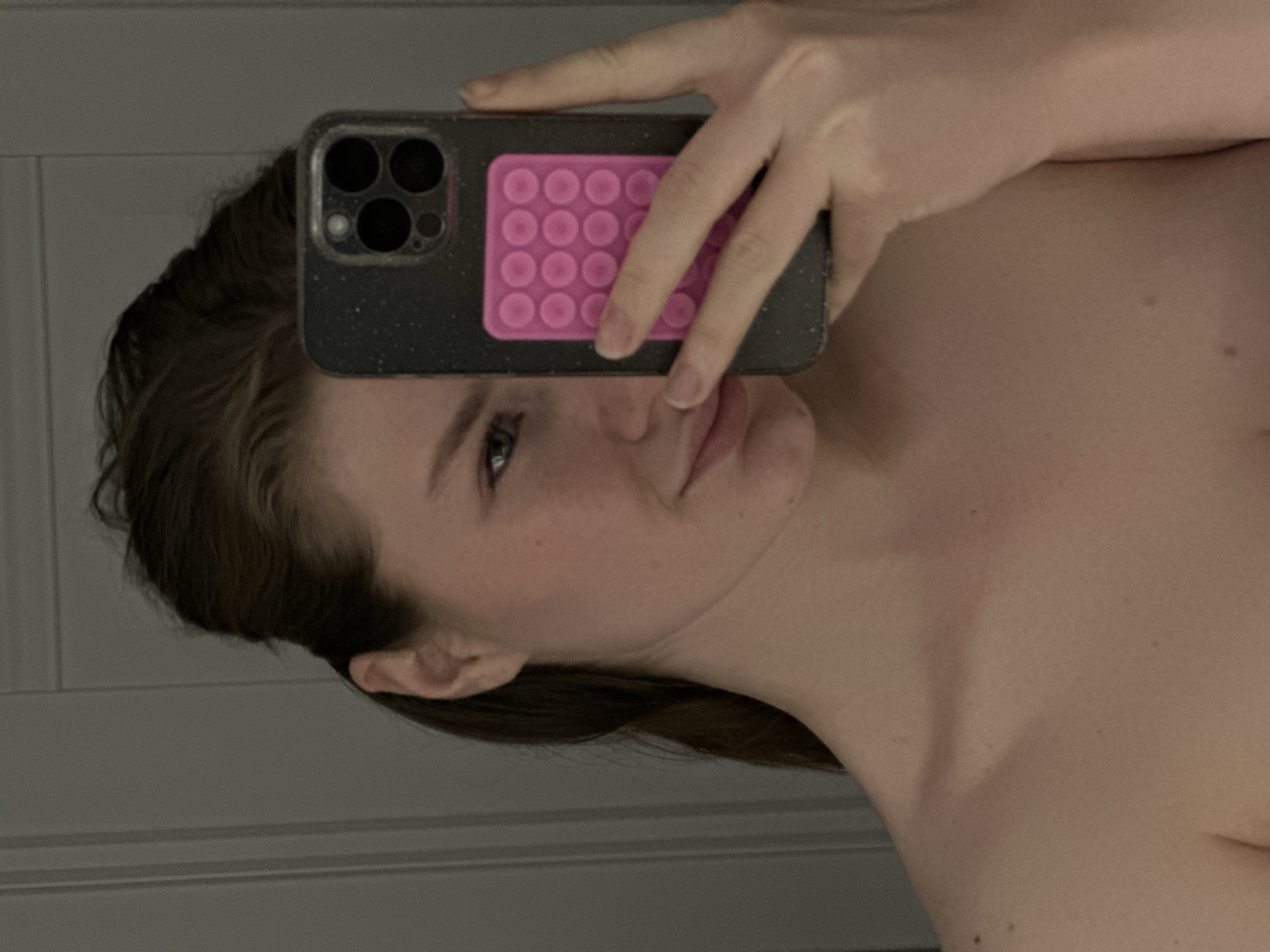 The image is a rotated mirror selfie featuring a person, likely a young woman, holding a smartphone. The main subject, a person with brown hair, occupies the right and lower portions of the frame, with their head to the right and their upper torso extending downwards. Their face is partially obscured by the phone, but one eye (the left eye), nose, and lips are visible, reflecting a neutral to slightly contemplative expression. Light freckles or moles are visible on their cheek and neck.
The person is holding a dark gray or black smartphone, which features a prominent camera module with three large dark lenses and a smaller light-colored circle (likely a flash or sensor), characteristic of certain iPhone models. The phone case has a speckled texture. Attached to the back of the phone case is a vibrant pink rectangular "pop-it" fidget toy, arranged in a 3x5 grid of 15 circular bubbles. The person's left hand is holding the phone, with fingers partially covering the pop-it, but the index finger and thumb are clearly visible gripping the device.
The background consists of a plain, light gray surface, possibly a wall or door, with subtle vertical lines suggesting paneling. The lighting is soft and even, creating a gentle illumination. The person appears to be bare-shouldered, with their upper chest and collarbone visible, implying they may be wearing an off-shoulder garment or are partially dressed. The overall color palette is subdued, with the bright pink pop-it providing a distinct color contrast.