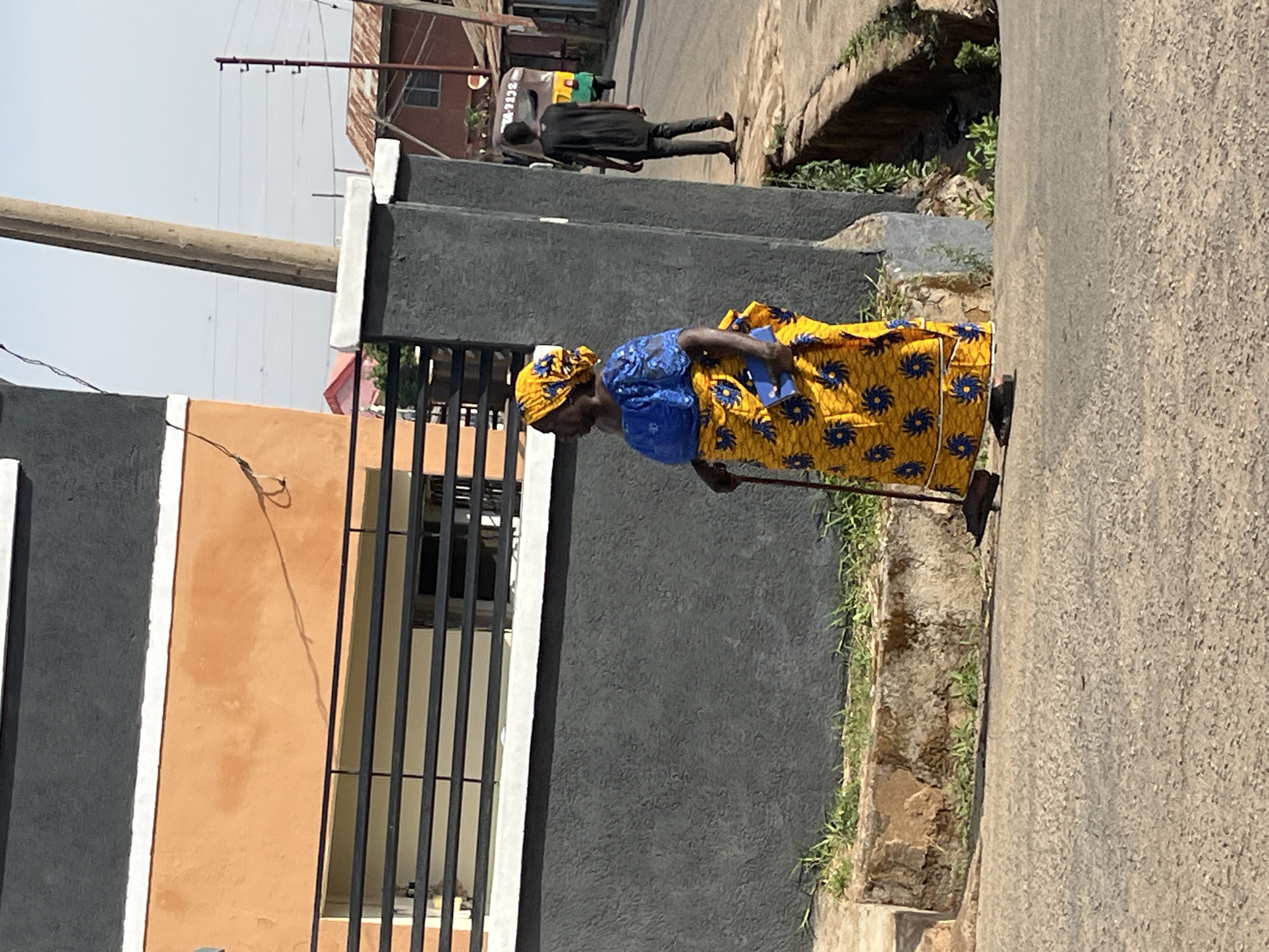 An elderly woman, attired in a yellow patterned dress and blue headscarf, is walking on a street in Jos, Nigeria. She uses a walking stick for support. A man in dark clothing is further up the road, and a three-wheeled vehicle can be seen in the background. The scene includes buildings, fences, and vegetation along the roadside.