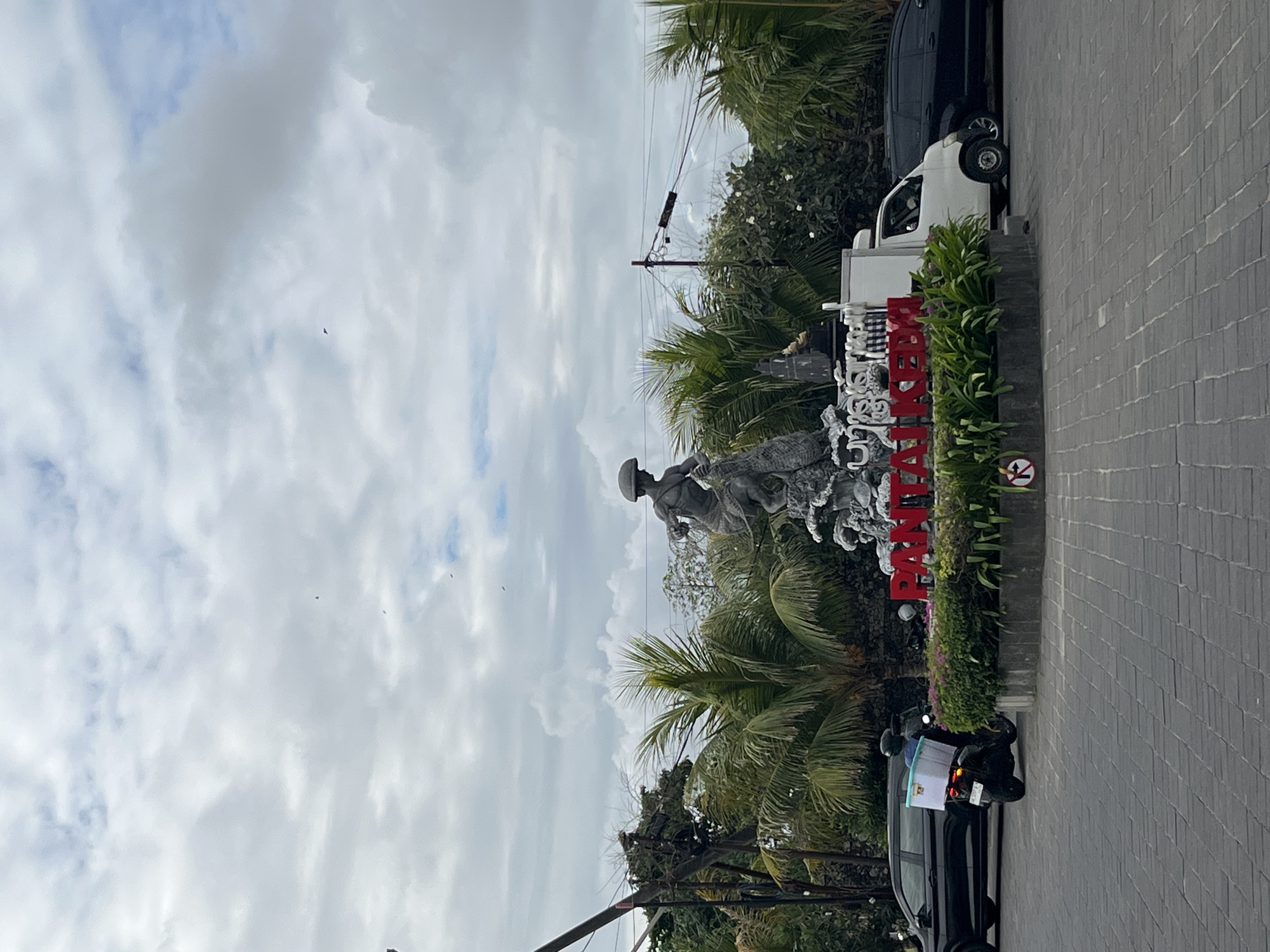 A statue depicting a fisherman is centrally located in a roundabout in Kuta, Bali, Indonesia. The statue is surrounded by lush greenery and palm trees. A large red sign with white letters that reads "PANTAI KUTA" is visible in front of the statue. Several vehicles are present on the road surrounding the roundabout, including a black car to the left, a white delivery van to the right, and a black car further to the right. A person on a scooter with a white cooler box attached to the back is seen in the foreground on the left. The sky is overcast with clouds.