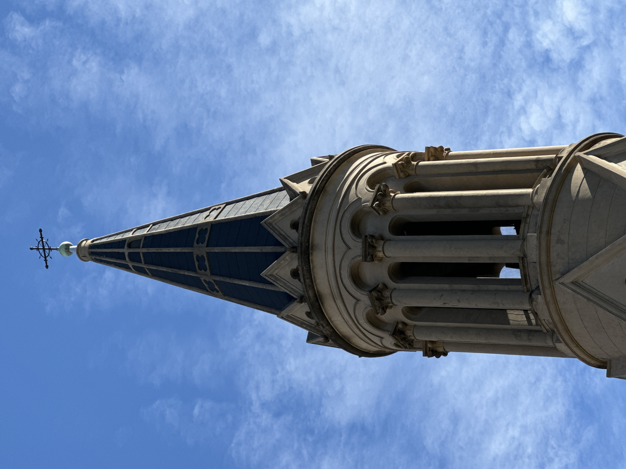 The image shows the top of a building spire against a blue sky with thin clouds. The spire has a cross at its peak, below which is a dark, patterned, sloping roof. Beneath the roof is a light-colored stone structure with pillars and ornamental detailing. The building is likely located in Buenos Aires, Argentina, based on architectural style.