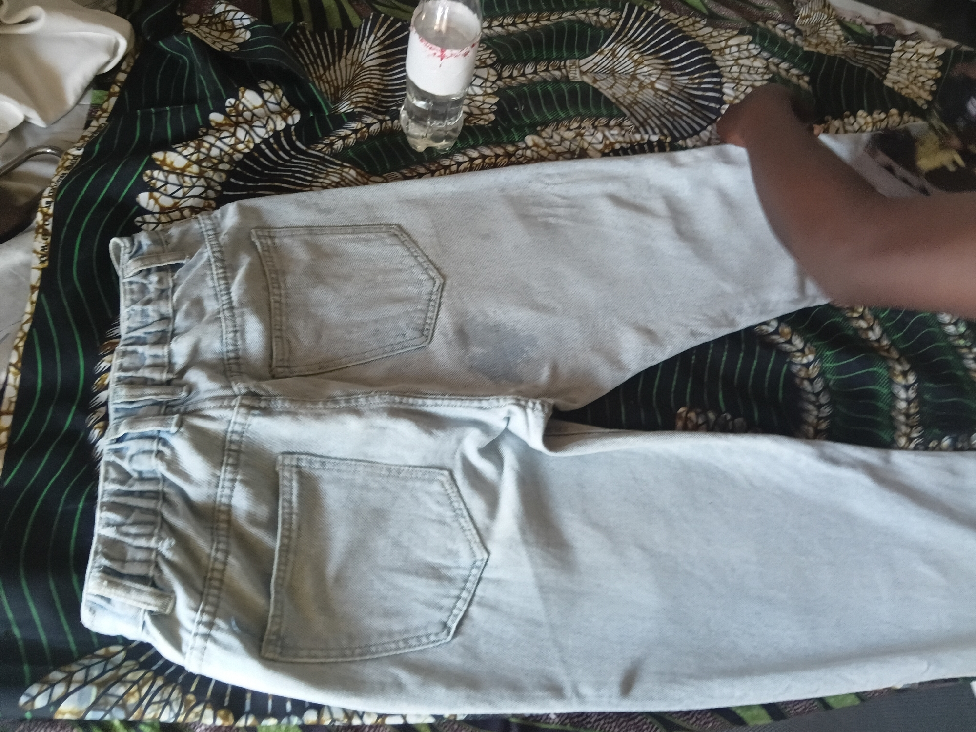 An overhead shot shows a pair of light blue denim jeans folded on top of a colorful fabric with a green, black, and white pattern. The jeans are laid out flat with the back pockets visible. One of the jeans legs is closer to the camera, showing the elastic waistband and a rear pocket. A clear plastic water bottle with a white label is positioned on the patterned fabric to the upper right of the jeans. The scene appears to be indoors, with the patterned fabric likely serving as a tablecloth or bedsheet. The lighting is natural, suggesting daylight. The image is focused on the jeans and fabric, with a person's arm and shoulder visible in the upper right corner, reaching towards the folded garment. There are no visible landmarks or contextual cues to determine the exact location within Jalingo, Nigeria.