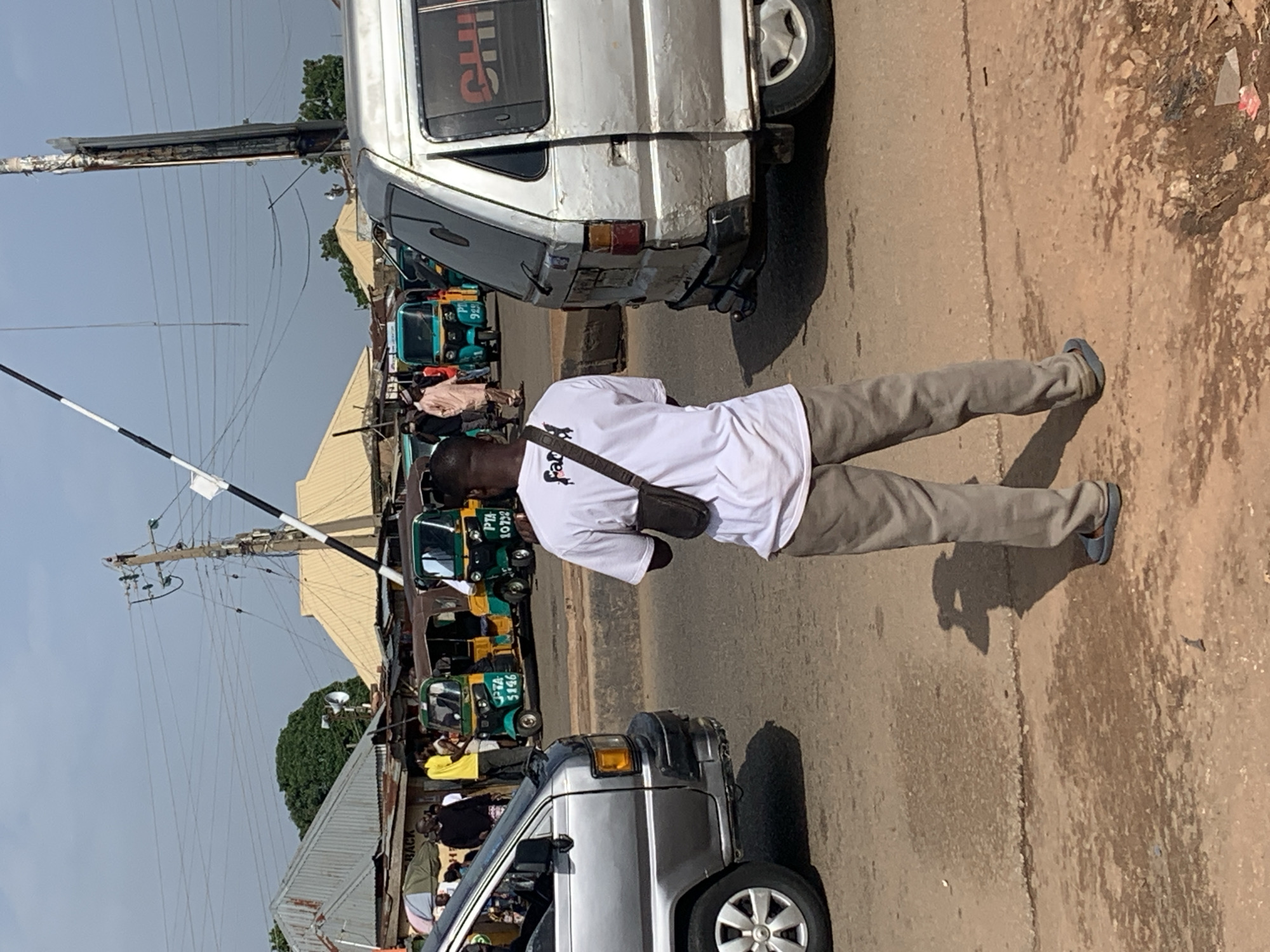 The image depicts a street scene in Gura Top, Nigeria, likely at a roadside market or intersection. In the foreground, a person wearing a white t-shirt with a dark graphic and grey pants stands with their back to the camera, looking towards a street with traffic. They are wearing a dark crossbody bag. To the left of this person is a silver van, and to the right is another silver van. Behind the vans, several colorful auto-rickshaws are visible, some with passengers and drivers. In the background, buildings with corrugated metal roofs line the street, and there are power lines and utility poles overhead. The overall environment appears to be a busy urban area with moderate traffic and pedestrian activity.
