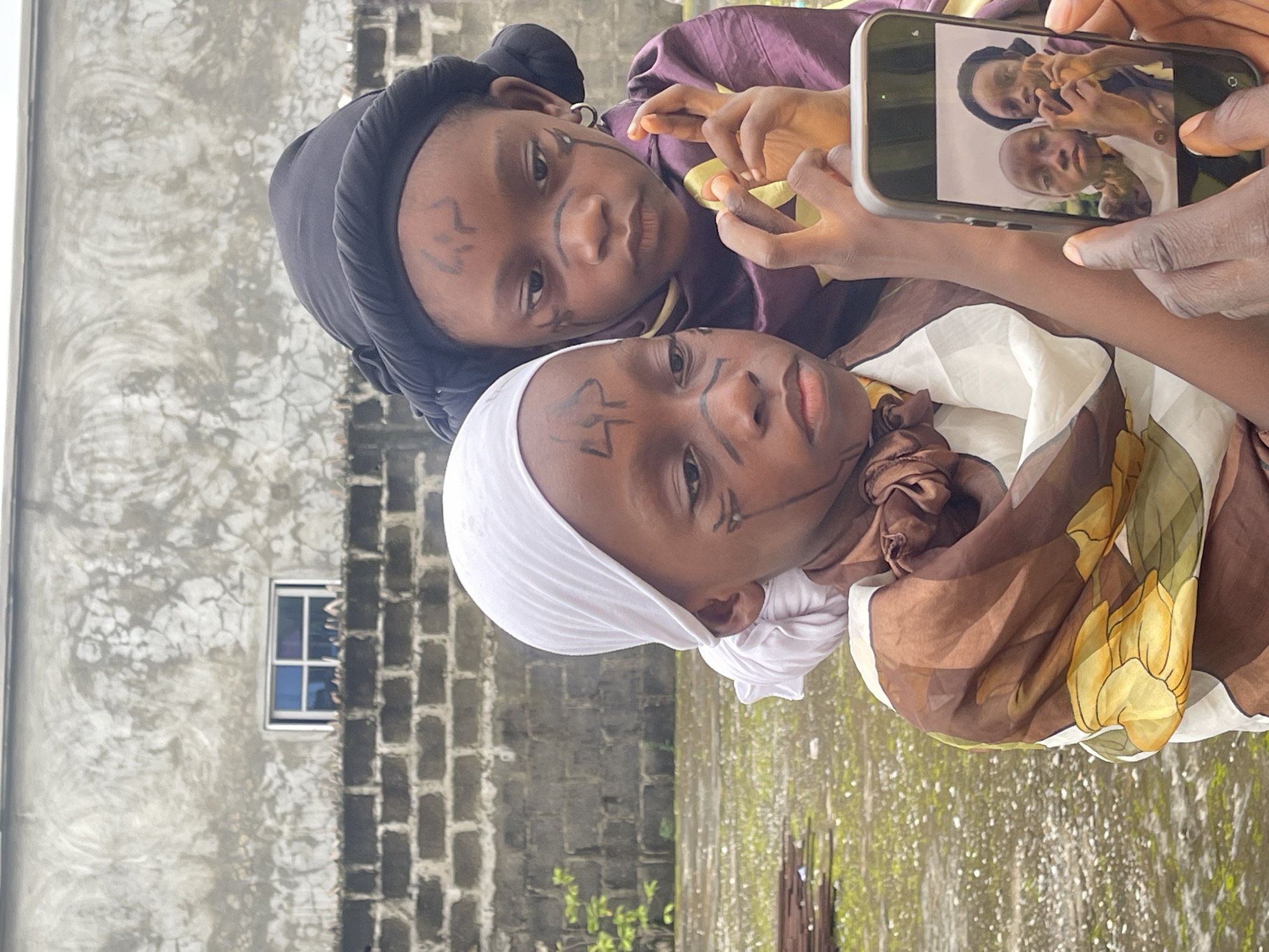 In Ado, Nigeria, two young children are featured in the foreground, posing for a selfie with a smartphone. The child on the left wears a white headscarf and a brown and yellow patterned garment, with black face paint on their forehead and cheeks. The child on the right is dressed in a dark headscarf and a purple garment, also with black face paint. They hold a smartphone displaying a photo of themselves, taken moments before.
The background consists of a weathered, grey concrete wall with a small, multi-pane window. Below the wall, a line of grey bricks is visible. The ground is wet and appears to be covered in gravel, with some green vegetation and metal rods in the distance. The overall scene suggests an outdoor setting, possibly in a less developed area of the city. The lighting indicates an overcast or cloudy day.