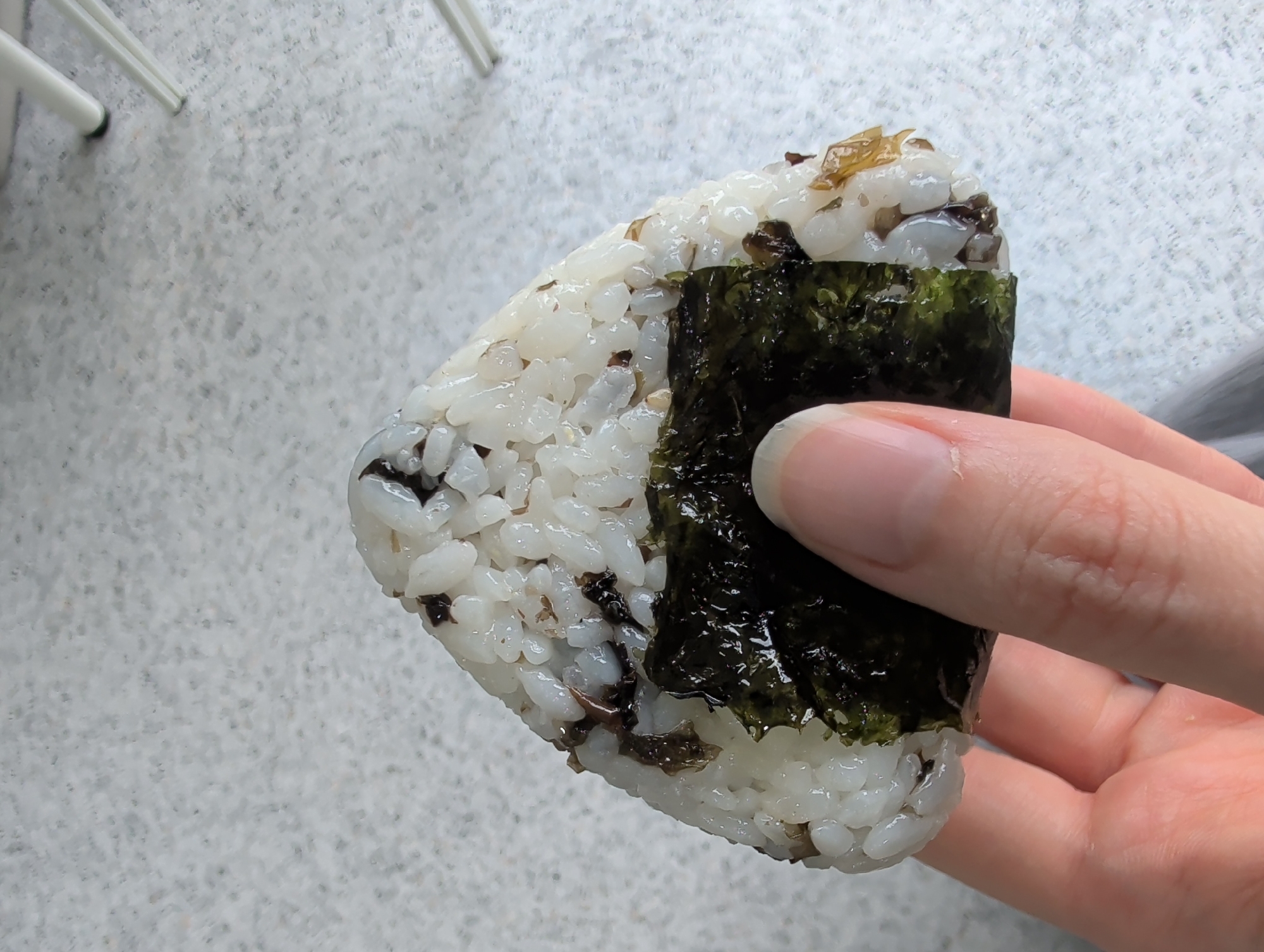 A person's hand holds a triangular onigiri, a Japanese rice ball wrapped with nori seaweed. The onigiri is primarily white rice with visible specks of black or dark brown ingredients mixed throughout. The seaweed is dark green and shiny. The hand holding the onigiri has fair skin and short fingernails. The background is a light gray speckled surface. A portion of white chair legs are visible in the upper left corner. The photo was likely taken indoors under natural light. The location is unknown.