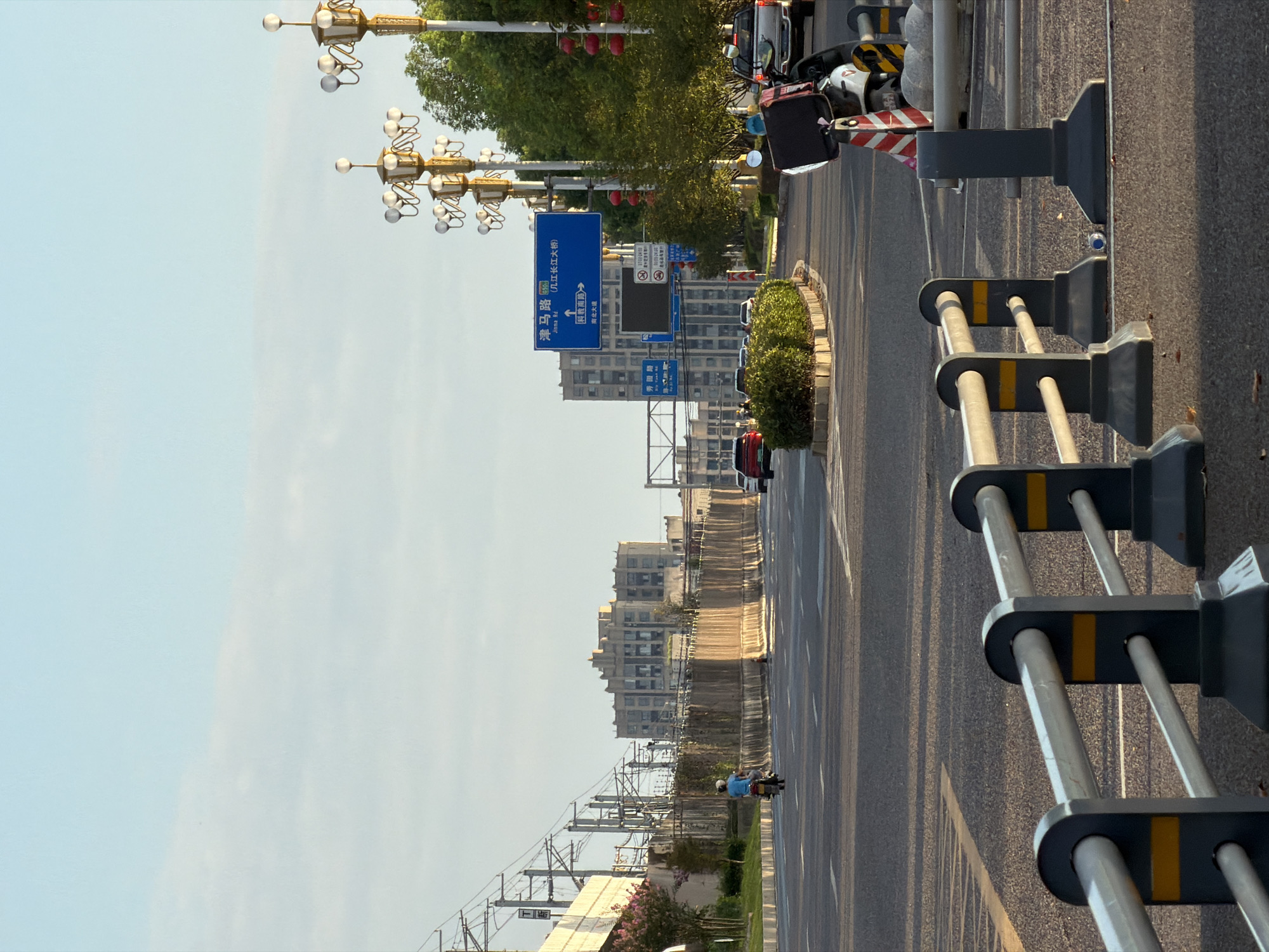 A street scene in Jiangjin, China, features a wide asphalt road with lane markings and reflective barriers. A single motorcyclist wearing a blue shirt and helmet travels away from the camera down the right lane. In the background, a multi-story building with a complex architectural design stands against a light blue sky with wispy clouds. To the left, an elevated structure with power lines suggests nearby train tracks. Traffic lights and street lamps with multiple globes are visible, some adorned with red lanterns. Blue road signs indicate directional information, including one with "Jinan Rd" and Chinese characters. Lush green trees line the right side of the road, and a circular raised planter with green bushes is positioned in the median. Several vehicles, including a silver pickup truck, are parked or in motion on the road. The sun casts long shadows, indicating either early morning or late afternoon light.