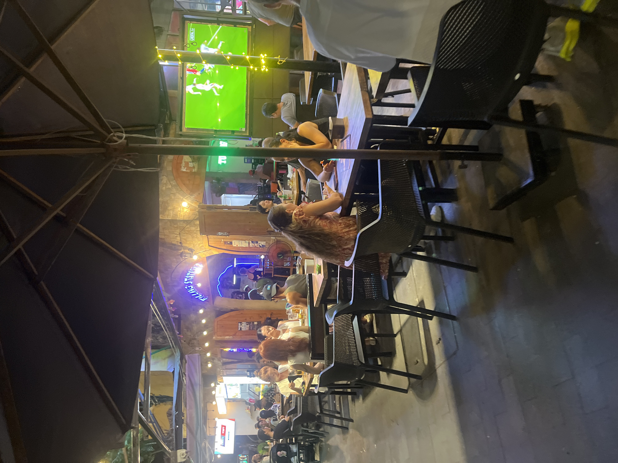 The image depicts an outdoor seating area of a bar or restaurant, likely in the city of Đà Nẵng, Vietnam, given the presence of a sign reading "FILLING STATION" in neon blue letters. The scene is illuminated by string lights and ambient lighting from the establishments. A large, dark umbrella provides shade over some of the tables.
Several groups of people are seated at tables, engaged in conversation and enjoying drinks. In the foreground, a woman with long, reddish-brown hair is seated at a table, looking towards her left. To her right, a woman with dark hair is visible, also seated at a table. Another group of individuals, including a woman with long dark hair and a man wearing a dark sleeveless shirt and a cap, are seated at a table further back.
On a large television screen in the background, a soccer match is being broadcast. The environment suggests a lively evening atmosphere, with patrons enjoying the entertainment and socializing. The flooring appears to be paved with stone or concrete. The overall impression is one of a casual, social gathering place.