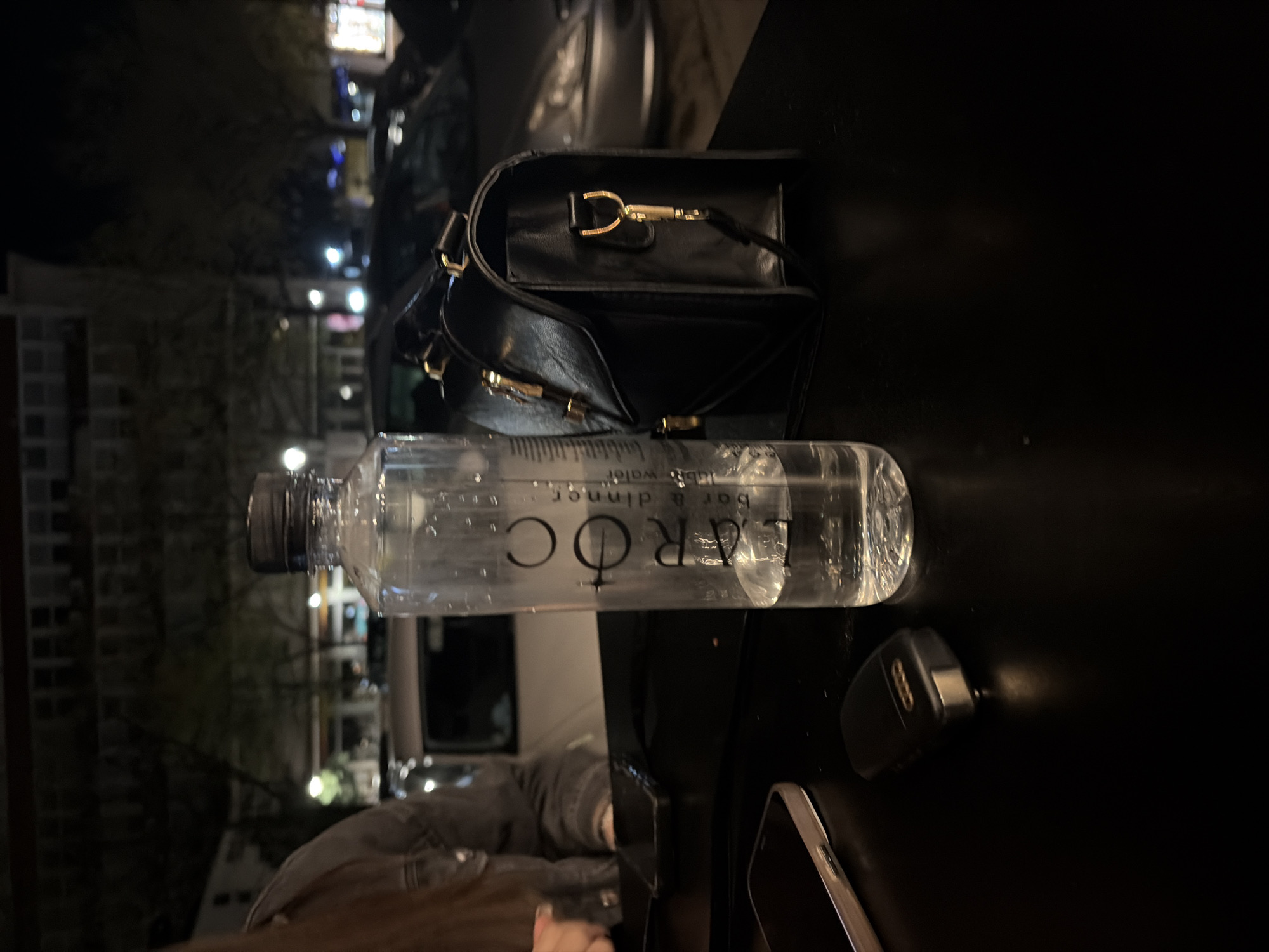 The image shows a close-up of a table surface at an outdoor location, likely a restaurant or bar. Dominating the scene is a plastic bottle of L'AROC water, partially full. Next to it is a black leather purse with gold hardware and a black car key fob with the Audi logo. A smartphone is also visible. The background features blurry city lights and buildings. Based on the information provided, this scene is at an outdoor setting somewhere in the city of Montana, Bulgaria.