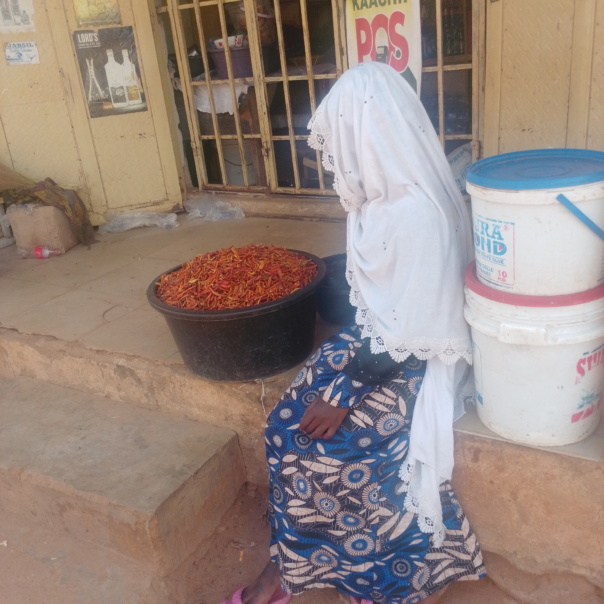 A person, likely a woman based on the attire, sits on steps in front of a store in Tempure, Nigeria. She is wearing a white headscarf with lace trim and a long blue and white patterned dress. To her left is a large black basin overflowing with dried red chilies. Behind her, stacked on the steps, are two large white buckets with blue and red lids. The store behind her has a metal grate across the entrance and several posters on the wall, one advertising "LORD'S CHOCOLATE GIN". Another sign above the grate reads "KAACHI POS". The ground is made of concrete and dirt. The lighting suggests it is daytime. The scene appears to be a market or street-side vendor.