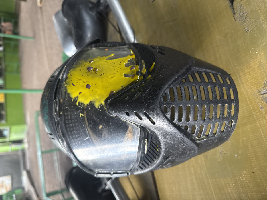 A black paintball mask sits with a splattering of yellow paint on its upper left side. The mask's visor is clear but partially obscured by the paint. Ventilation holes are visible on the right side. The background appears to be an outdoor setting, with glimpses of greenery and other equipment. The surface underneath the mask is tan and slightly worn.
