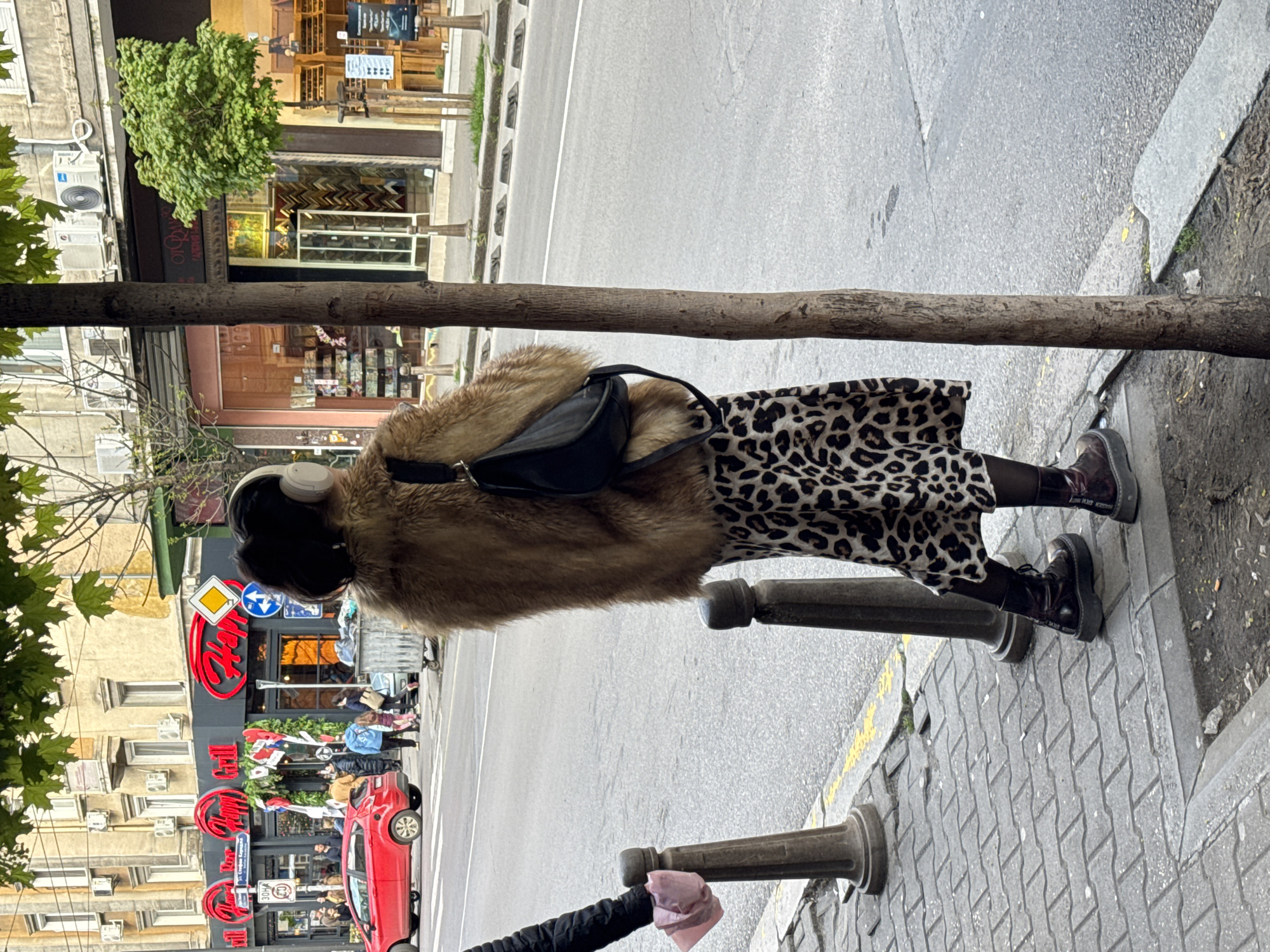 The image, oriented vertically, depicts a daytime urban street scene in what appears to be Sofia, Bulgaria. The primary subject is a person, seen from behind and slightly to the left, standing or walking on a paved sidewalk. This individual is wearing a brown, possibly faux-fur, coat, a black and white leopard print midi-skirt, dark tights or stockings, and chunky, dark boots with "RANGER STEVE" visible in white lettering on the side. White over-ear headphones are worn, and a black shoulder bag is slung over the right shoulder.
A prominent tree trunk extends horizontally across the mid-ground, segmenting the view. To the left of the tree, lining the street, are multi-story buildings with ground-floor shops and restaurants. Visible details include air conditioning units on facades, display windows (one appearing to show picture frames), and a prominent red sign with the word "Happy" in stylized lettering. Further down, other shops and pedestrians are visible.
To the right of the tree, the gray asphalt road is visible, featuring a white lane marking. Along the curb, two dark, cylindrical bollards are placed, with a small pink item resting on one in the foreground. A red car is seen in the distance on the road, amidst other pedestrians. Street signs, including a yellow diamond and a blue circular sign with arrows, are also present. The lighting is even, suggesting an overcast or partly cloudy day, characteristic of a typical urban street during daytime.