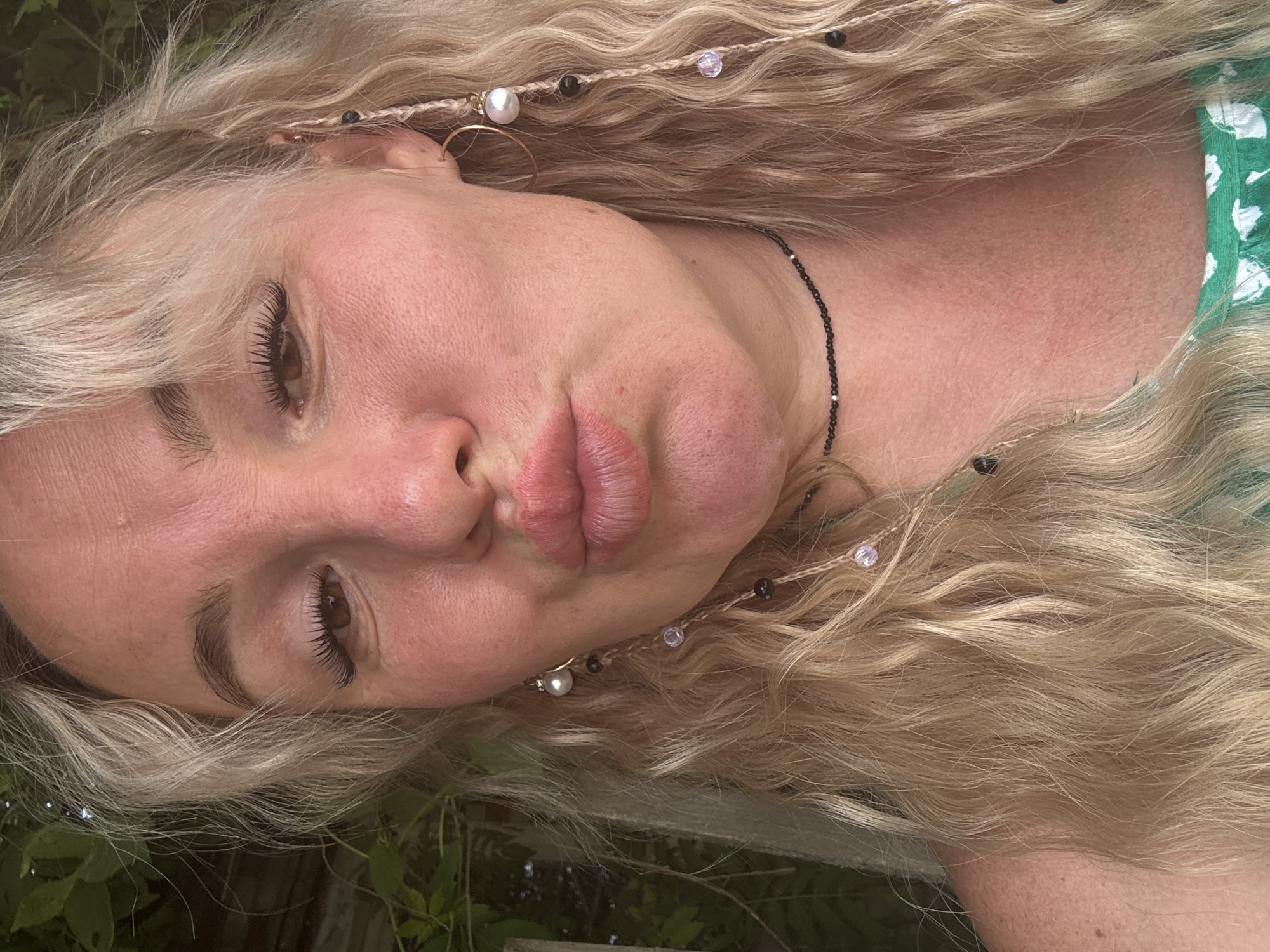 A close-up, outdoor selfie captures a fair-skinned woman with long, blonde, wavy hair and a tan complexion. Her face is slightly flushed, with light makeup enhancing her eyes and lips. She is puckering her lips, creating a kissing gesture, and her eyes are looking towards the camera. Her hair is adorned with braided strands featuring small beads and pearls. She wears a thin, black choker necklace with small metallic beads. The background is softly blurred, suggesting a natural, outdoor environment, possibly a garden with green foliage visible. The image appears to be taken in natural daylight.